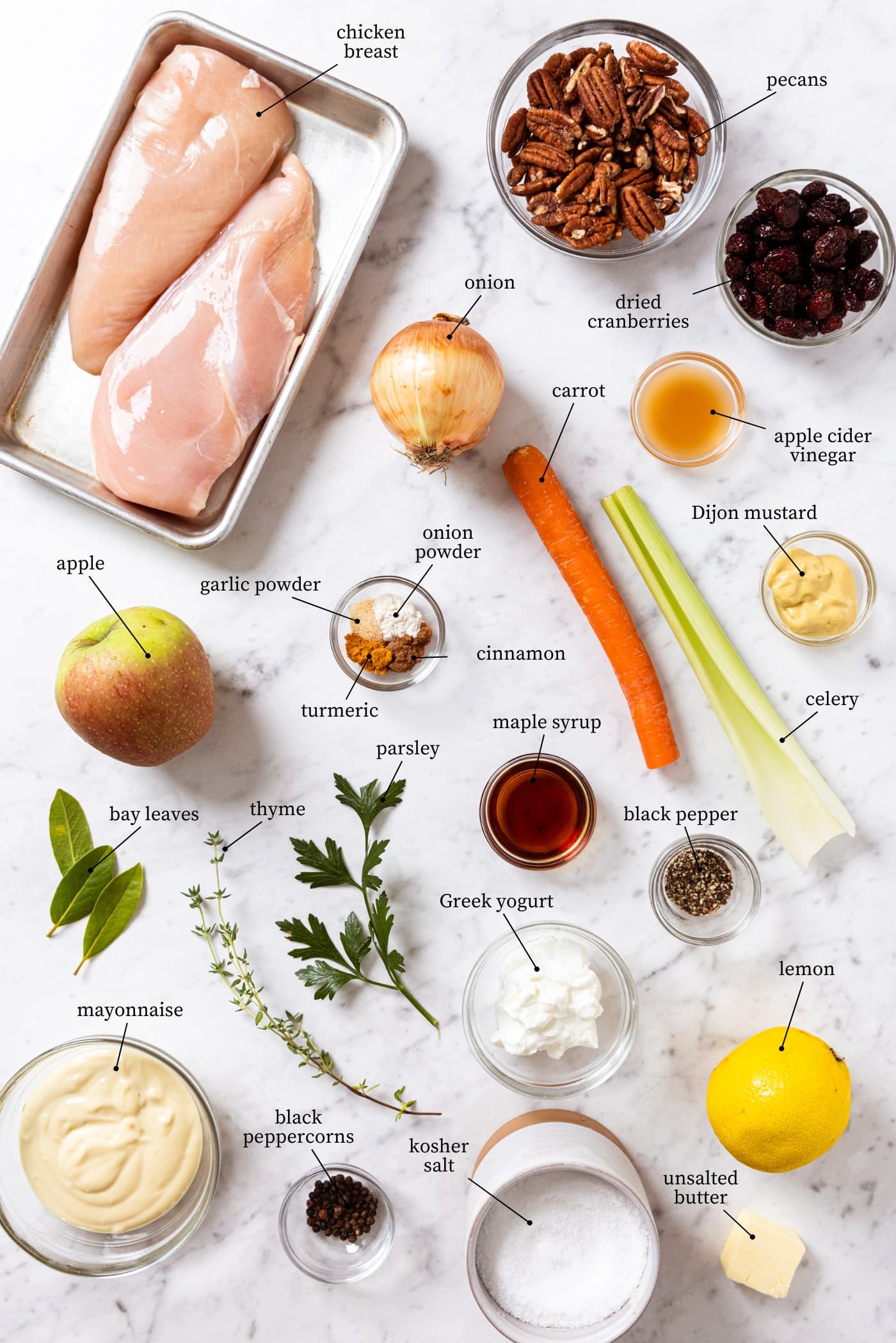 White marble surface filled with all ingredients needed to make chicken salad including raw chicken breasts, pecans, spices, mayonnaise, Greek yogurt, and more.