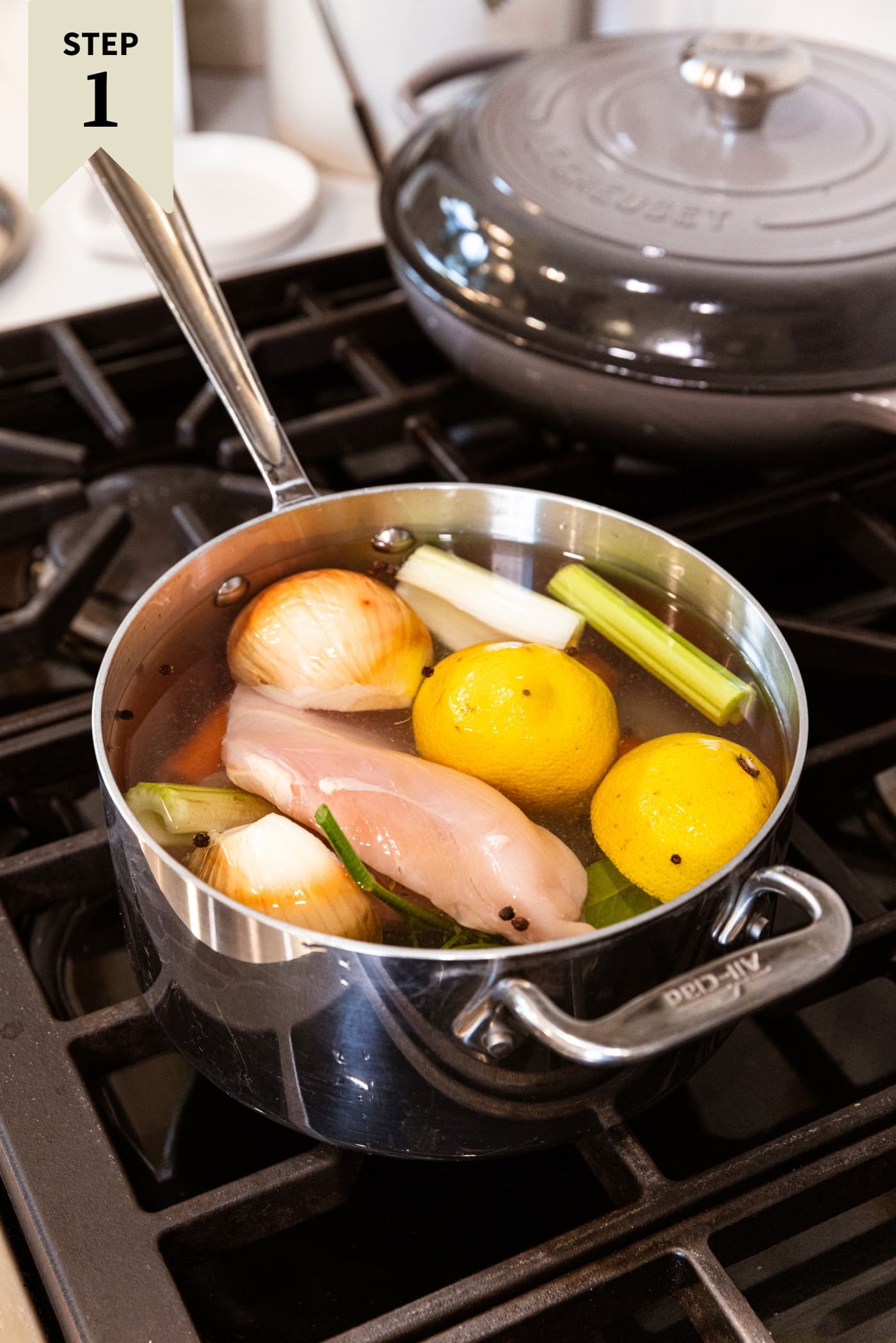 Large metal kettle filled with raw chicken breasts ready to poach in water with lemon, celery, carrot, and onion.