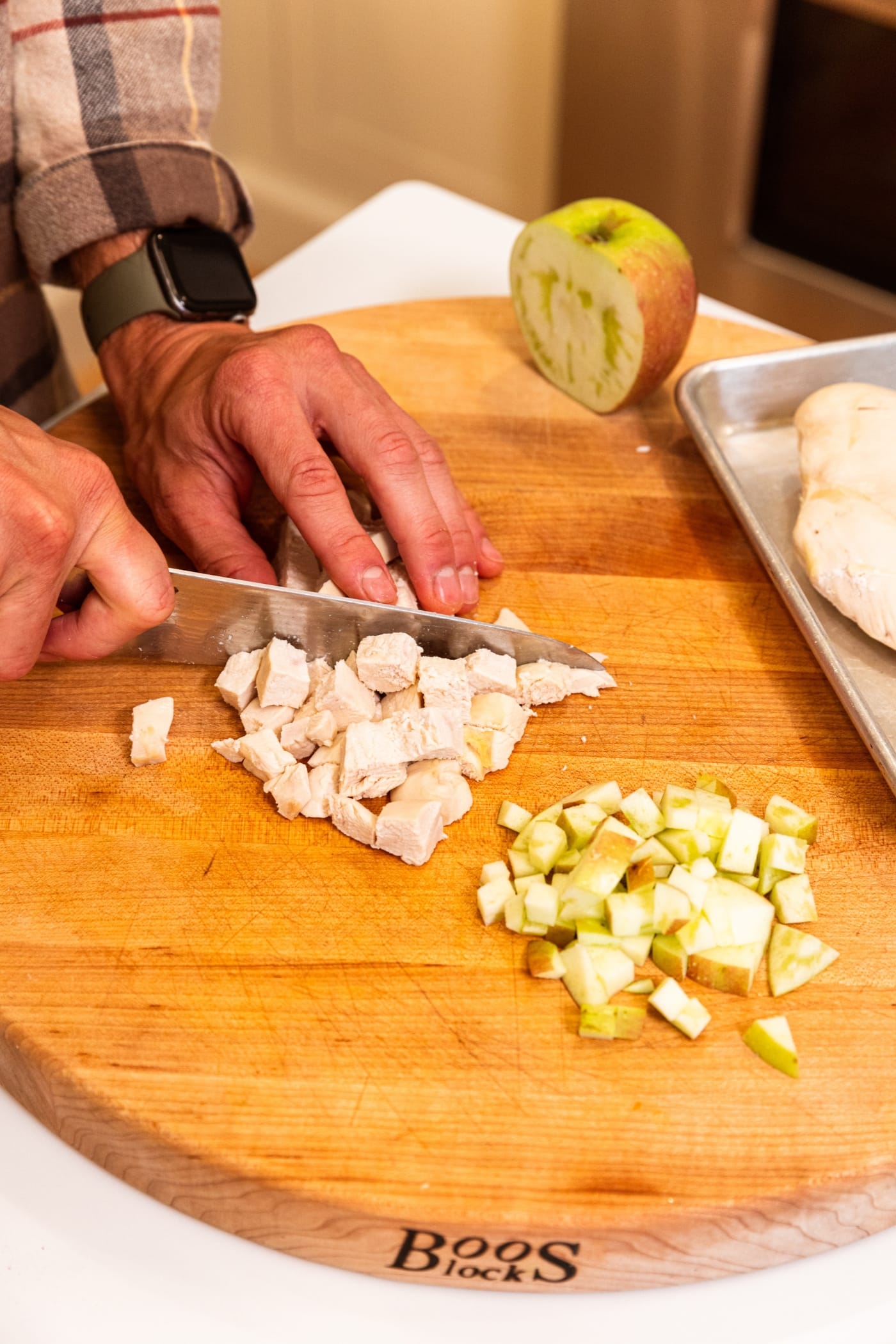 Two hands using knife to slice poached chicken into small cubes along with some apple.