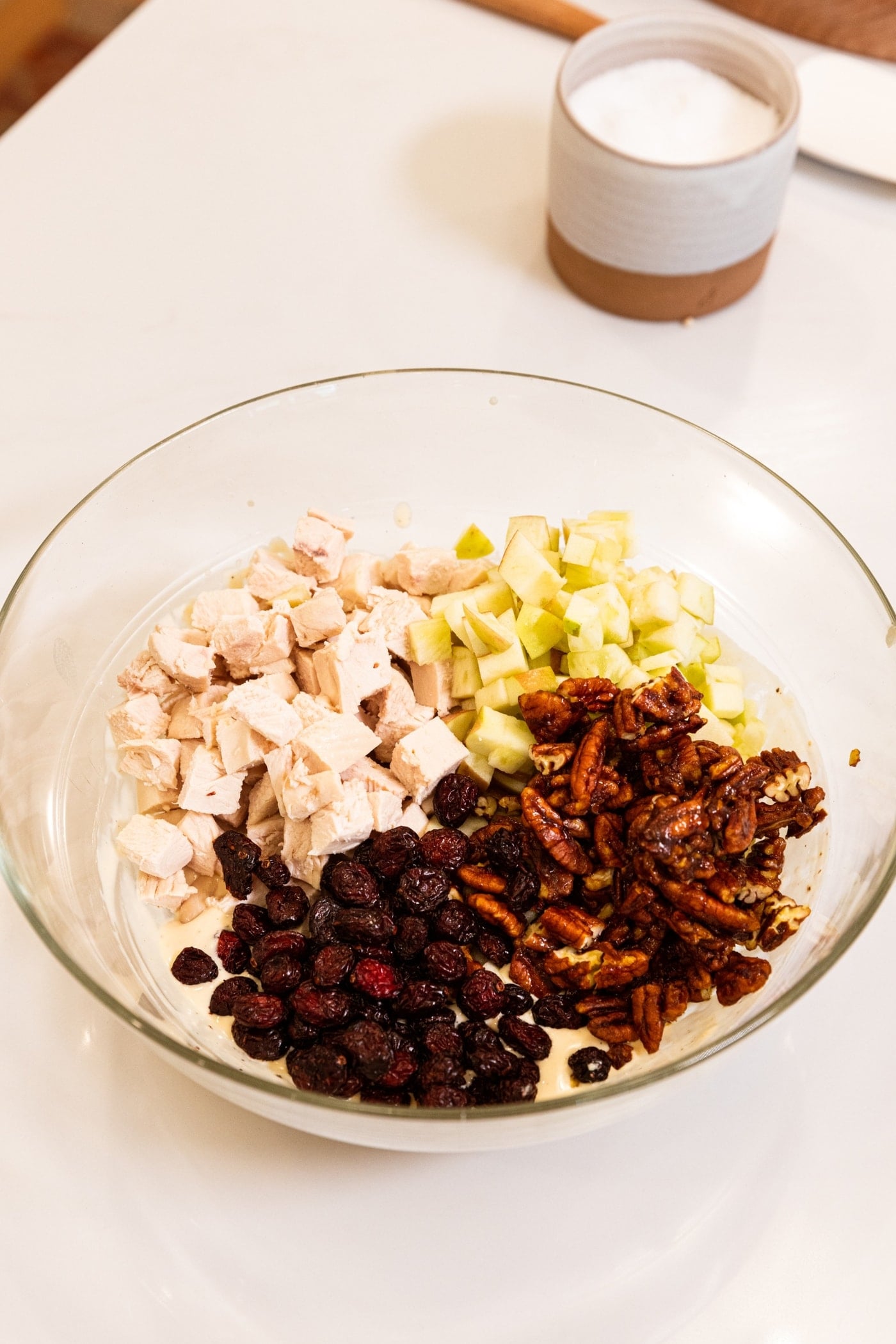Large glass bowl filled with chopped poached chicken, dried cranberries, candied pecans, and diced apples.
