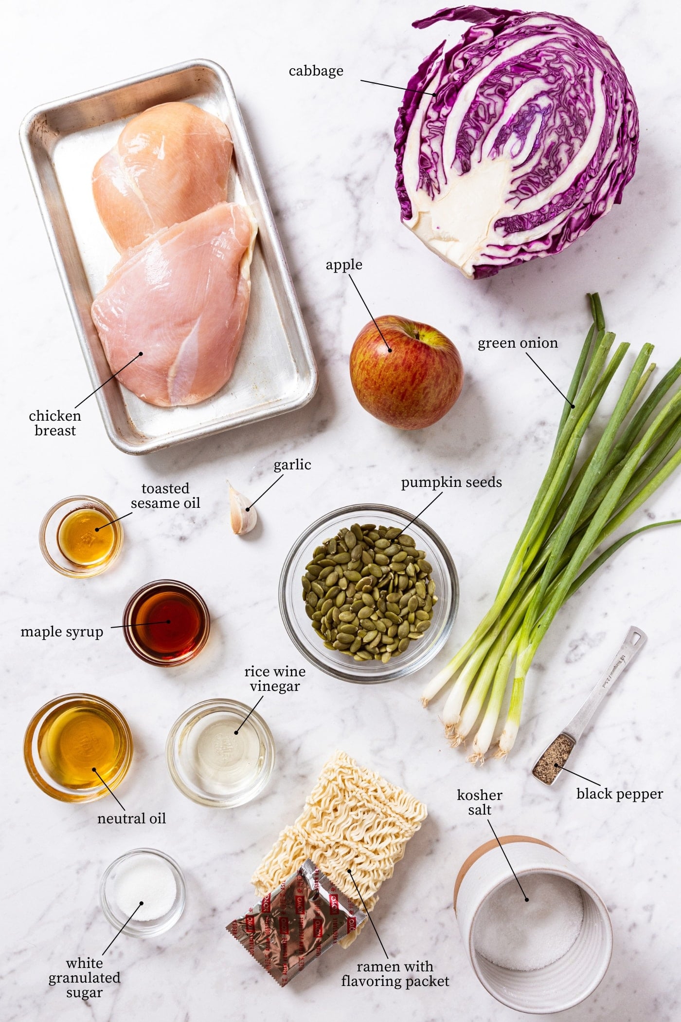 White marble surface filled with all ingredients needed to make ramen chicken salad including chicken breasts, ramen noodles, cabbage, green onions, oils, spices, and more.