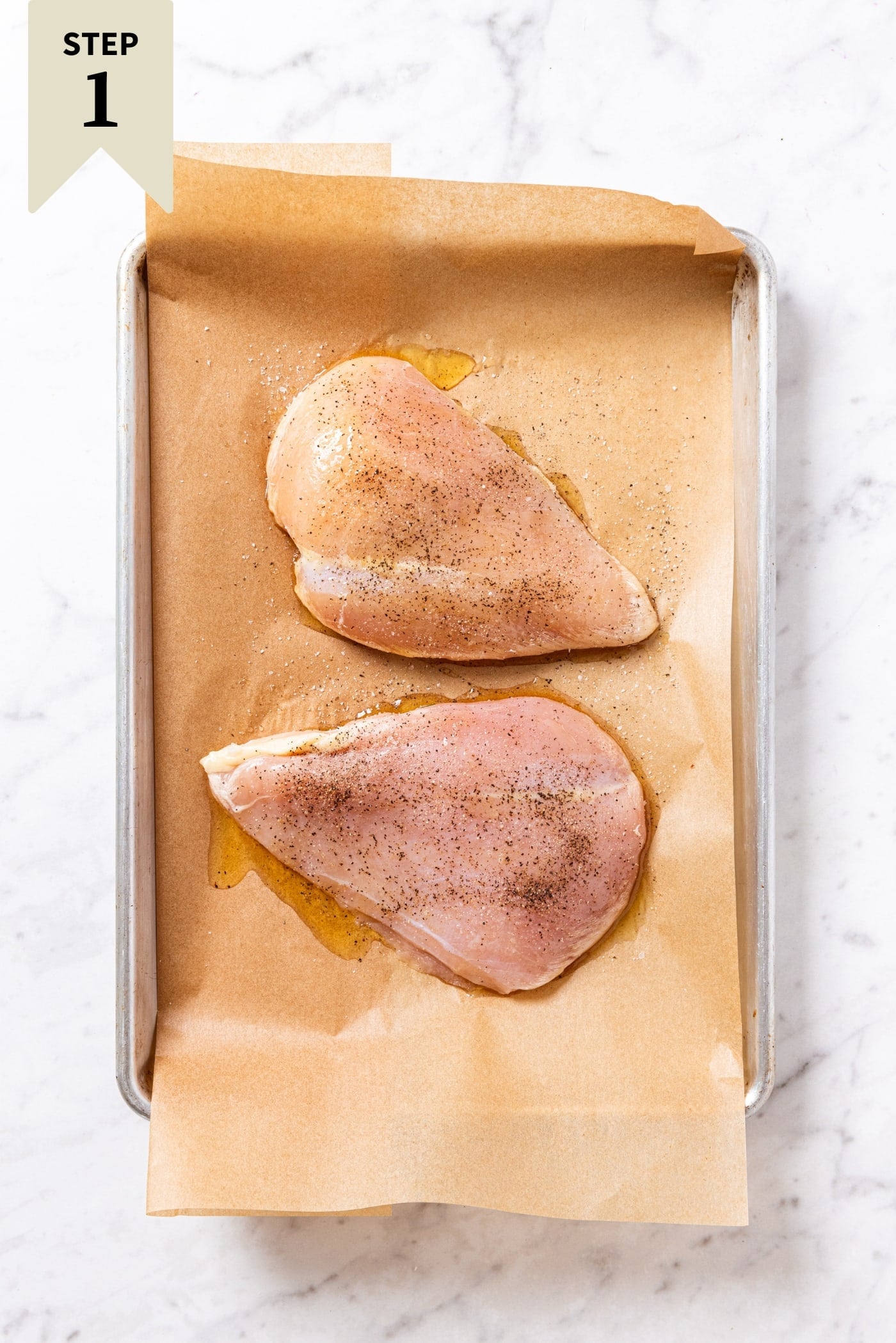 Two pieces of raw chicken breast sitting on piece of parchment paper in a baking sheet on white marble before going into the oven.