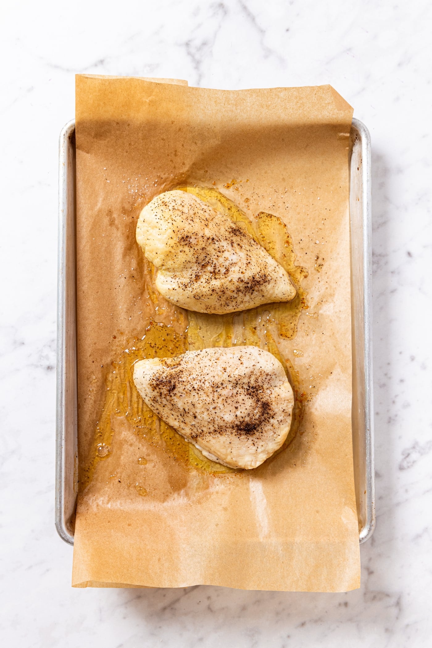 Two chicken breasts after coming out of the oven with oil and salt and pepper all over on piece of parchment paper.