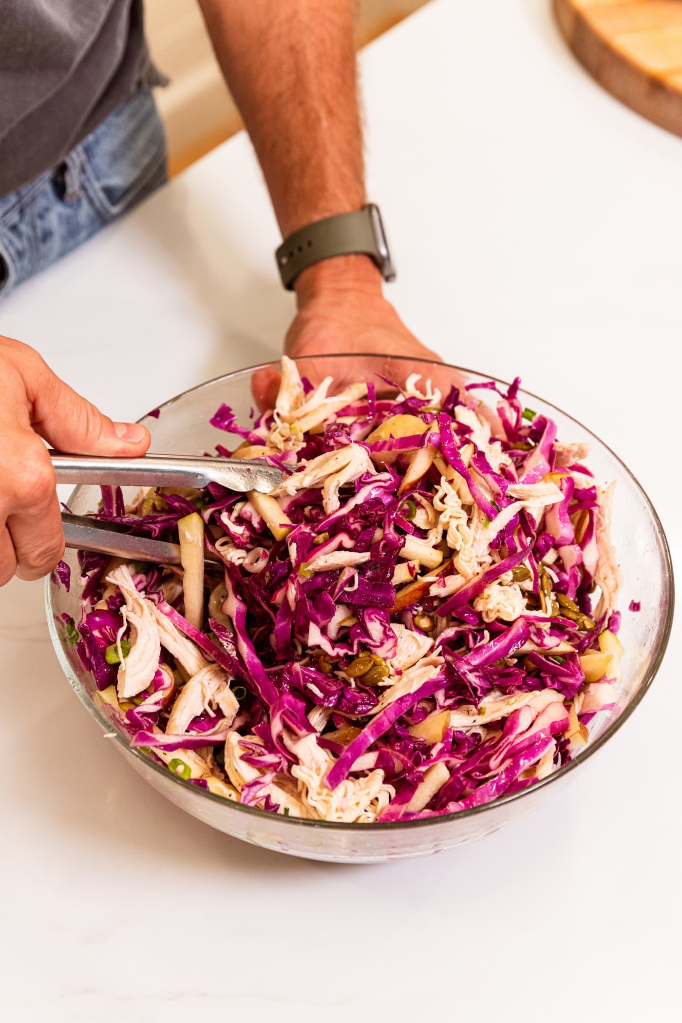 Hand using tongs to toss together all components of ramen chicken salad with red cabbage.