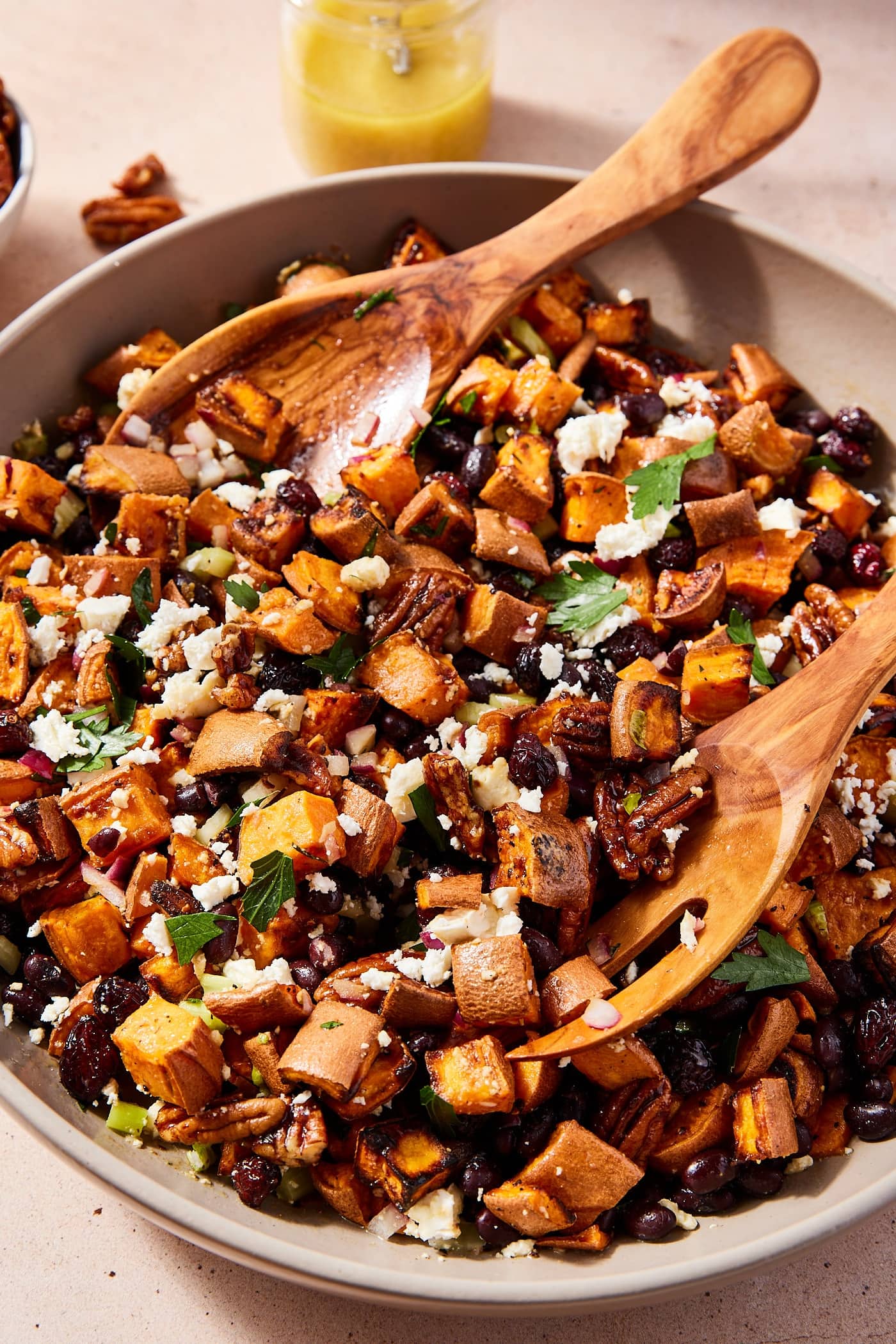 Two wooden tongs resting on side of brown bowl after mixing together roasted sweet potato salad topped with herbs and feta cheese.