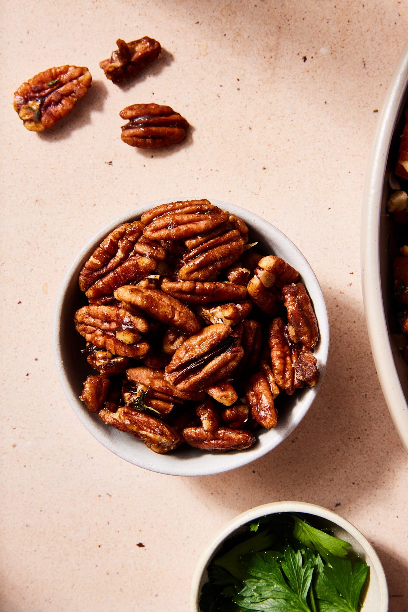 Small bowl filled with candied pecan halves with a few on the table beside the bowl along with fresh herbs.