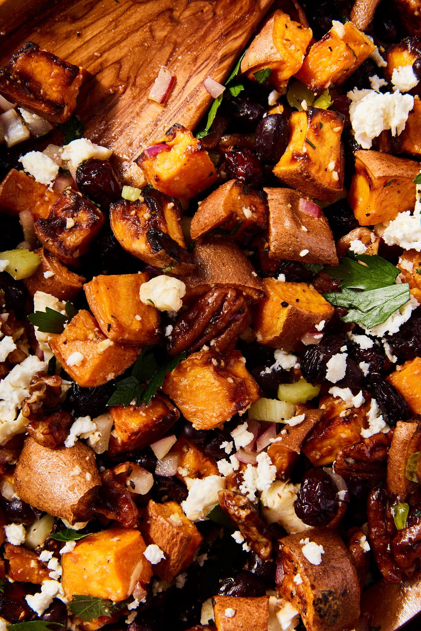 Mixture of roasted sweet potatoes with feta cheese and herbs all in a bowl with wooden tongs mixing everything together.