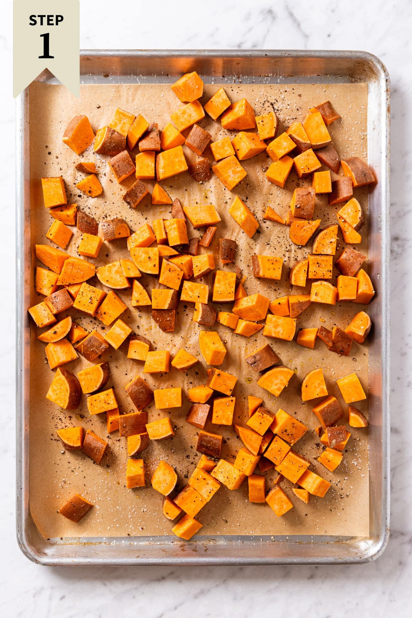 Uncooked raw sweet potato pieces on parchment-lined baking sheet on white marble surface right before going into the oven to bake.