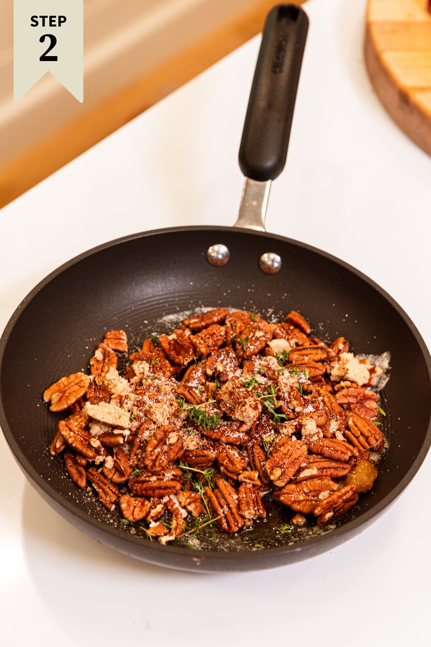 Skillet resting on a white surface filled with pecans, spices, and some fresh herbs before being cooked on the stove.
