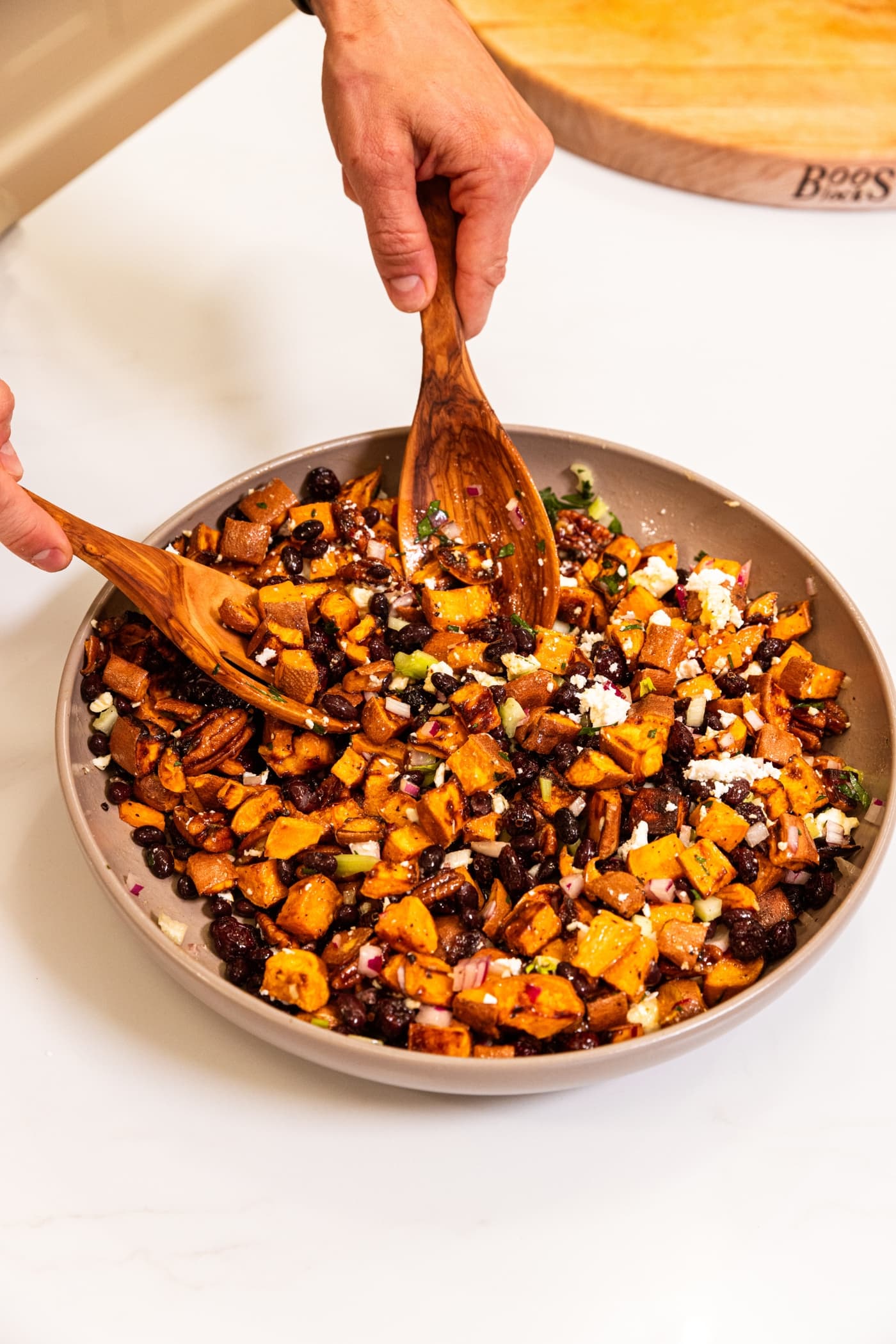 Two hand holding tongs mixing together sweet potato salad ingredients in a large brown bowl on white surface.