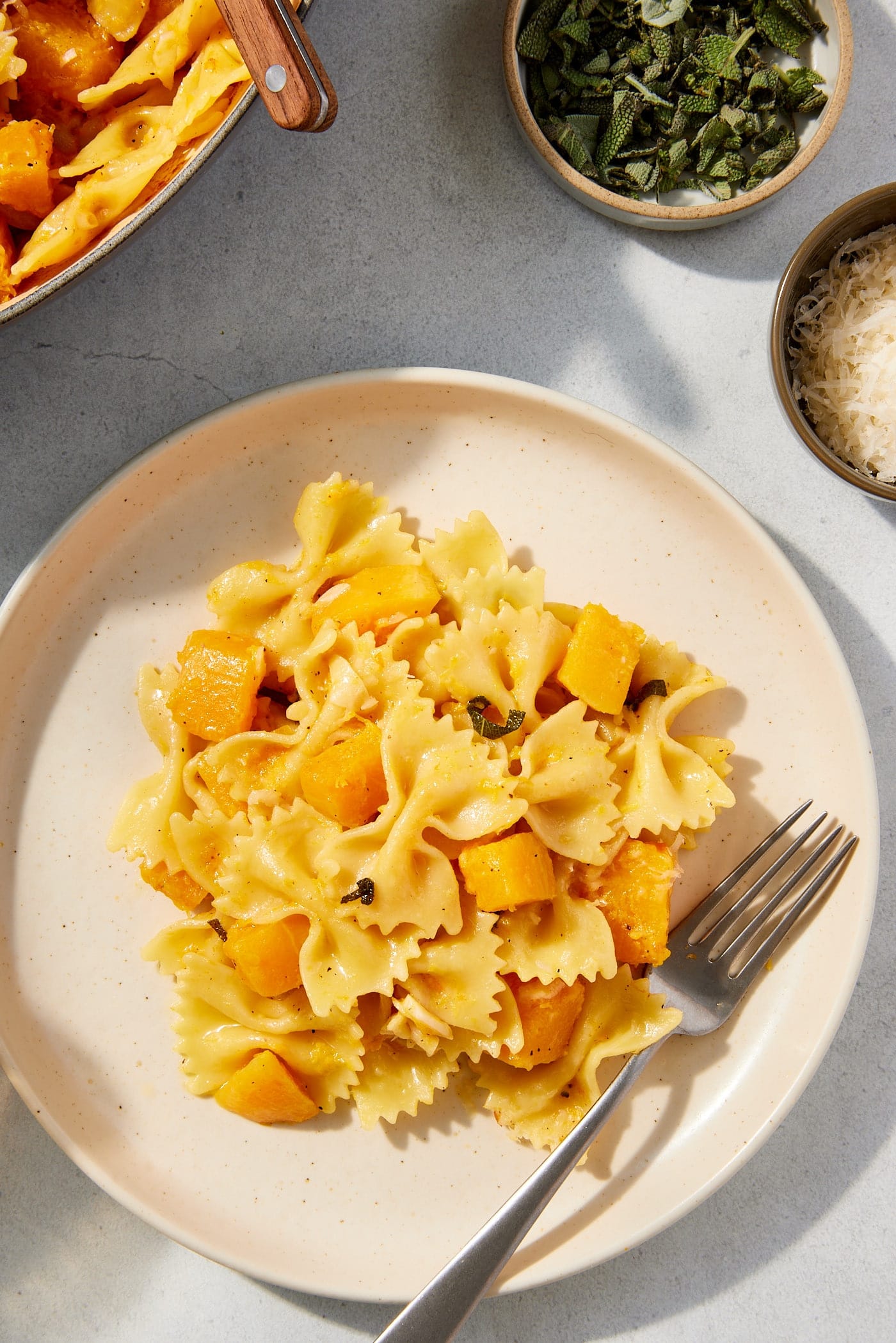 White plate filled with serving of browned butter squash pasta with fork resting to the side with small bowl filled with Parmesan cheese.