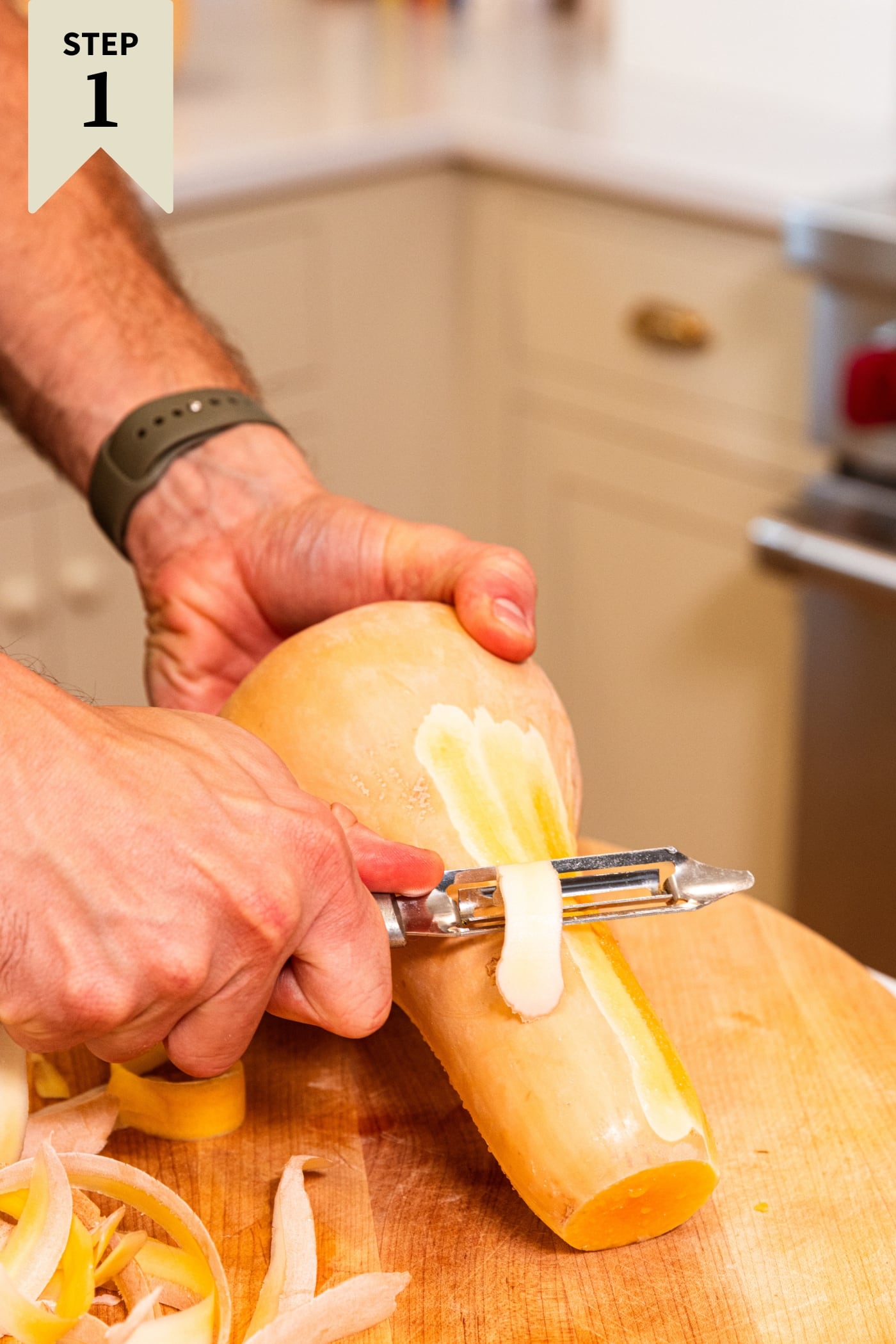 Hand holding butternut squash peeling it with a peeler on a wooden cutting board.