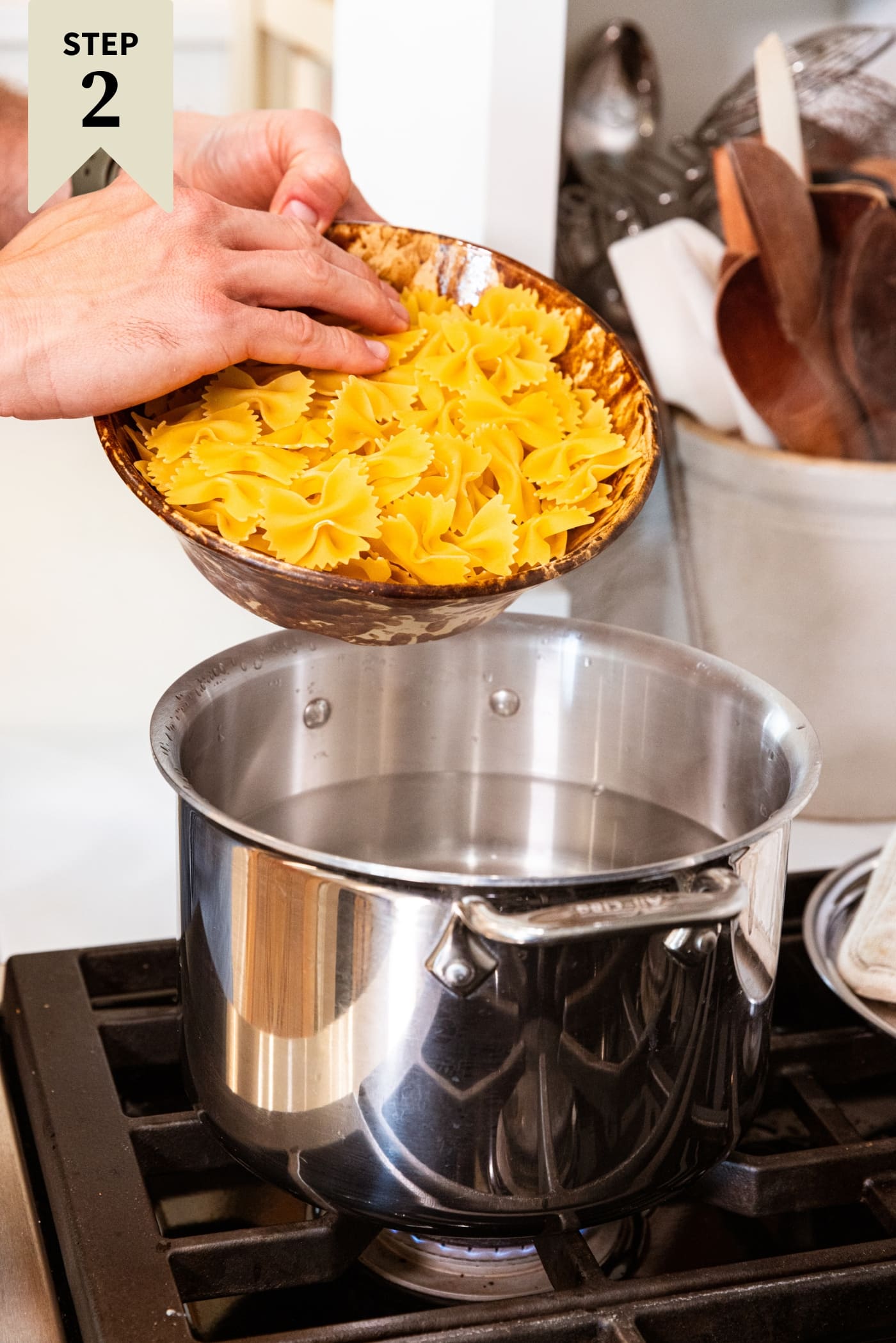 Hand holding brown bowl about to pour raw pasta into a large stockpot filled with water.