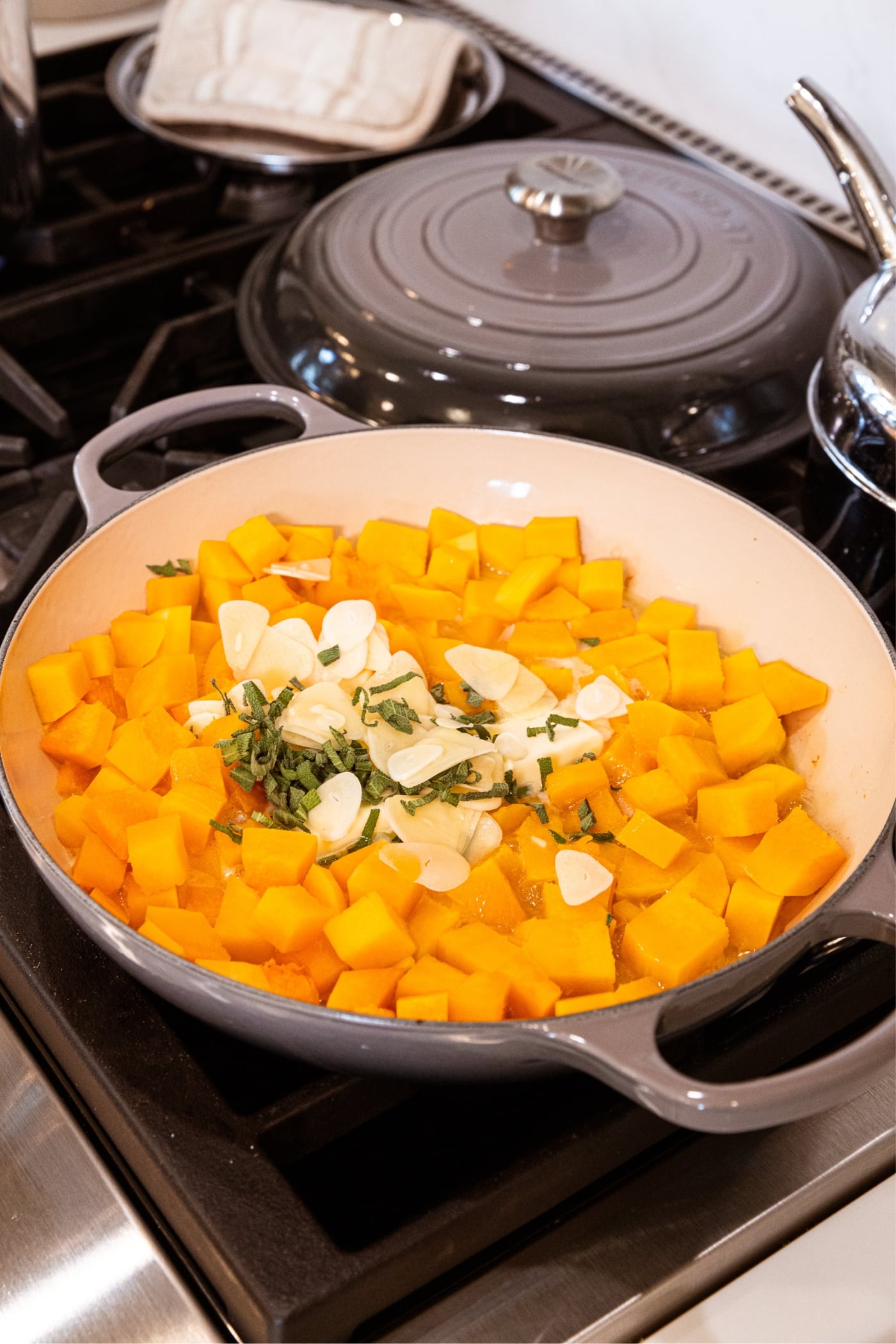 Large gray skillet filled with cooked butternut squash with chopped fresh herbs and sliced garlic cooking on stovetop.