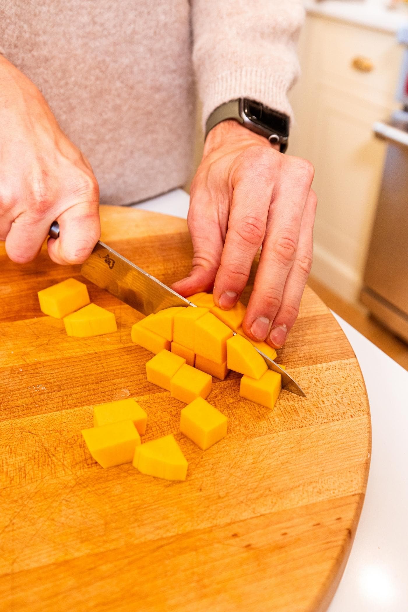 Two hands holding knife slicing butternut squash into cubes on wooden cutting board.