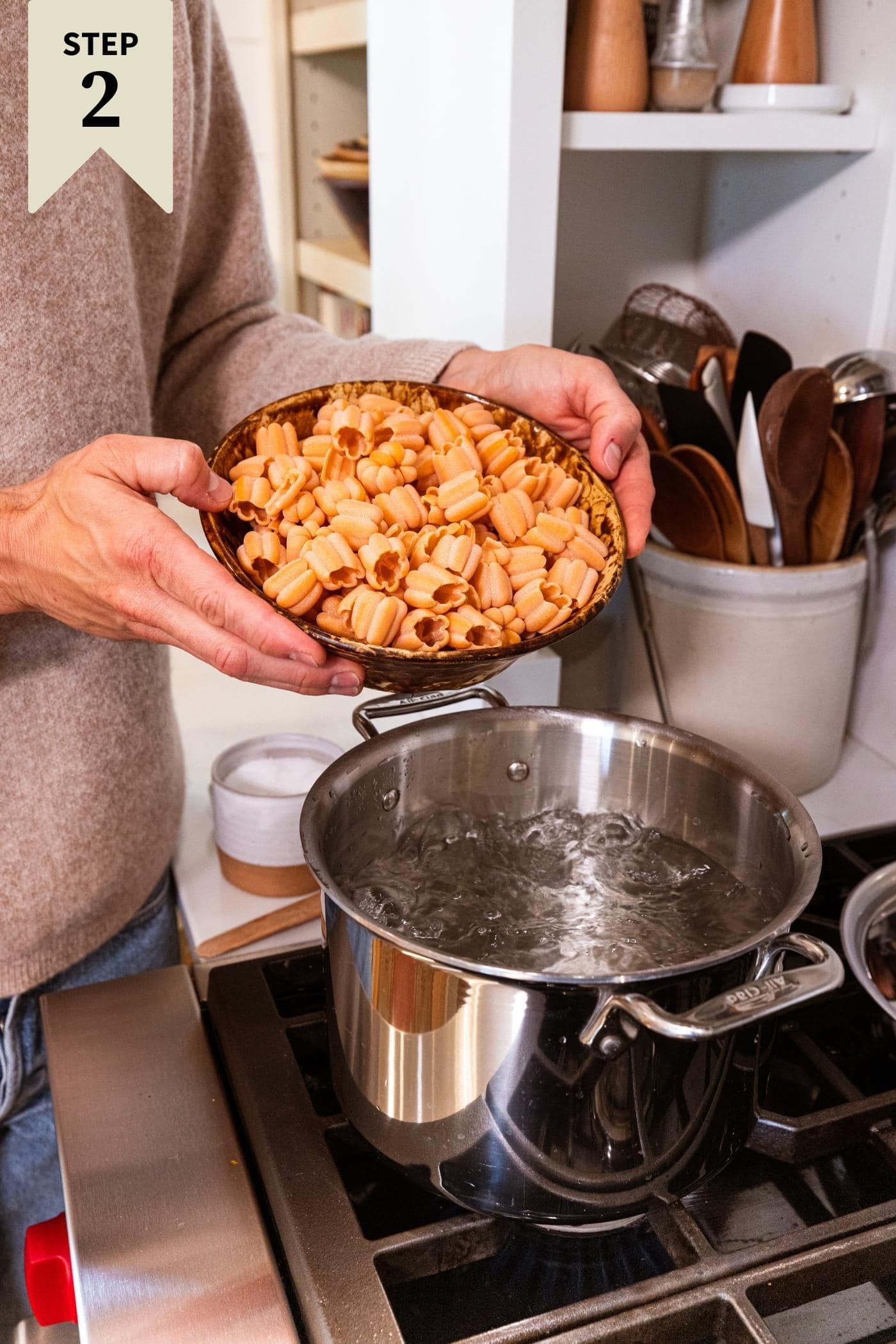 Two hands pouring brown colored pasta into a large stockpot filled with boiling water on stovetop.