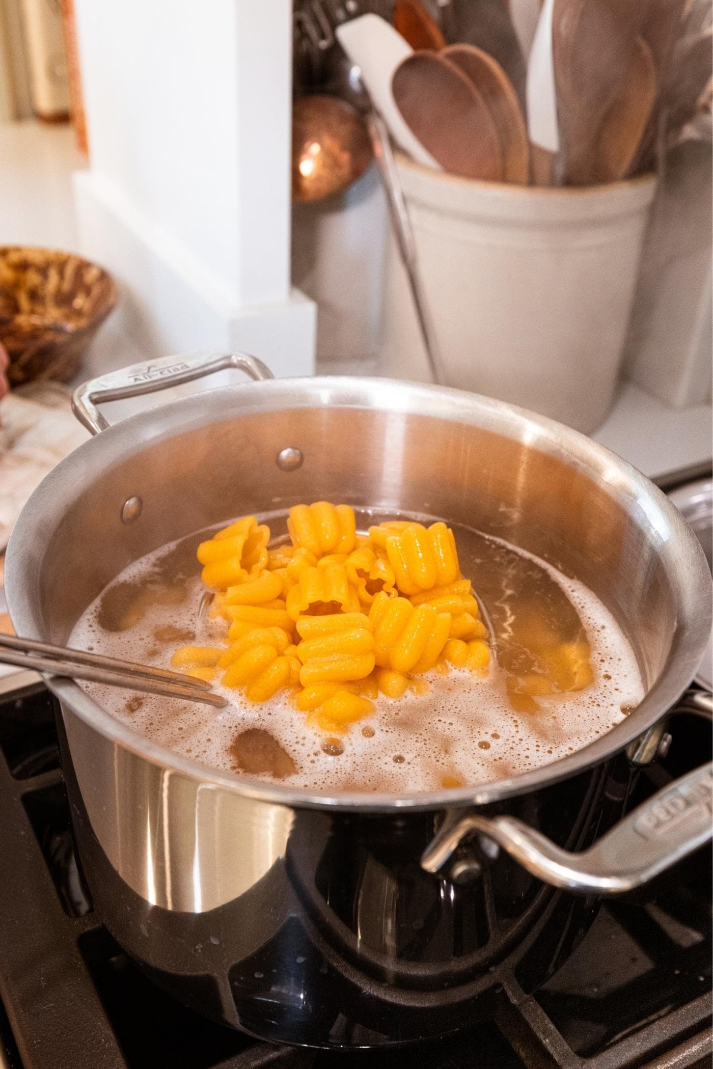 Hand holding spider scooping cooked pieces of pasta from stockpot filled with water on the stove.