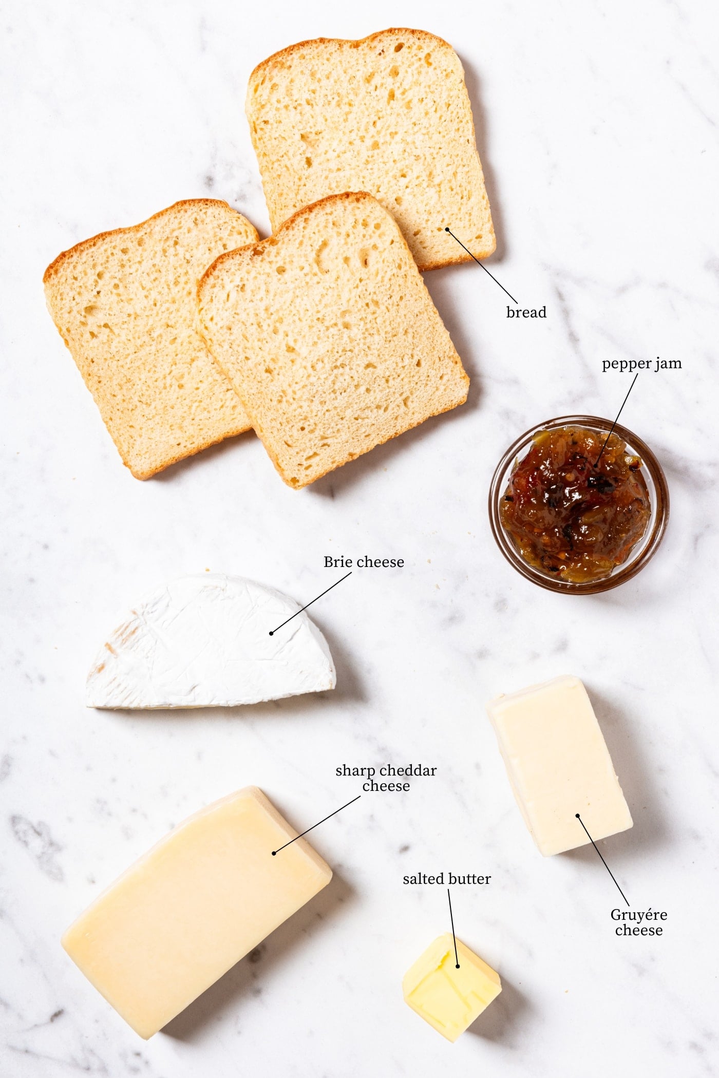White marble surface filled with all ingredients needed to make grilled cheese croutons including slices of bread, jam, and cheeses.
