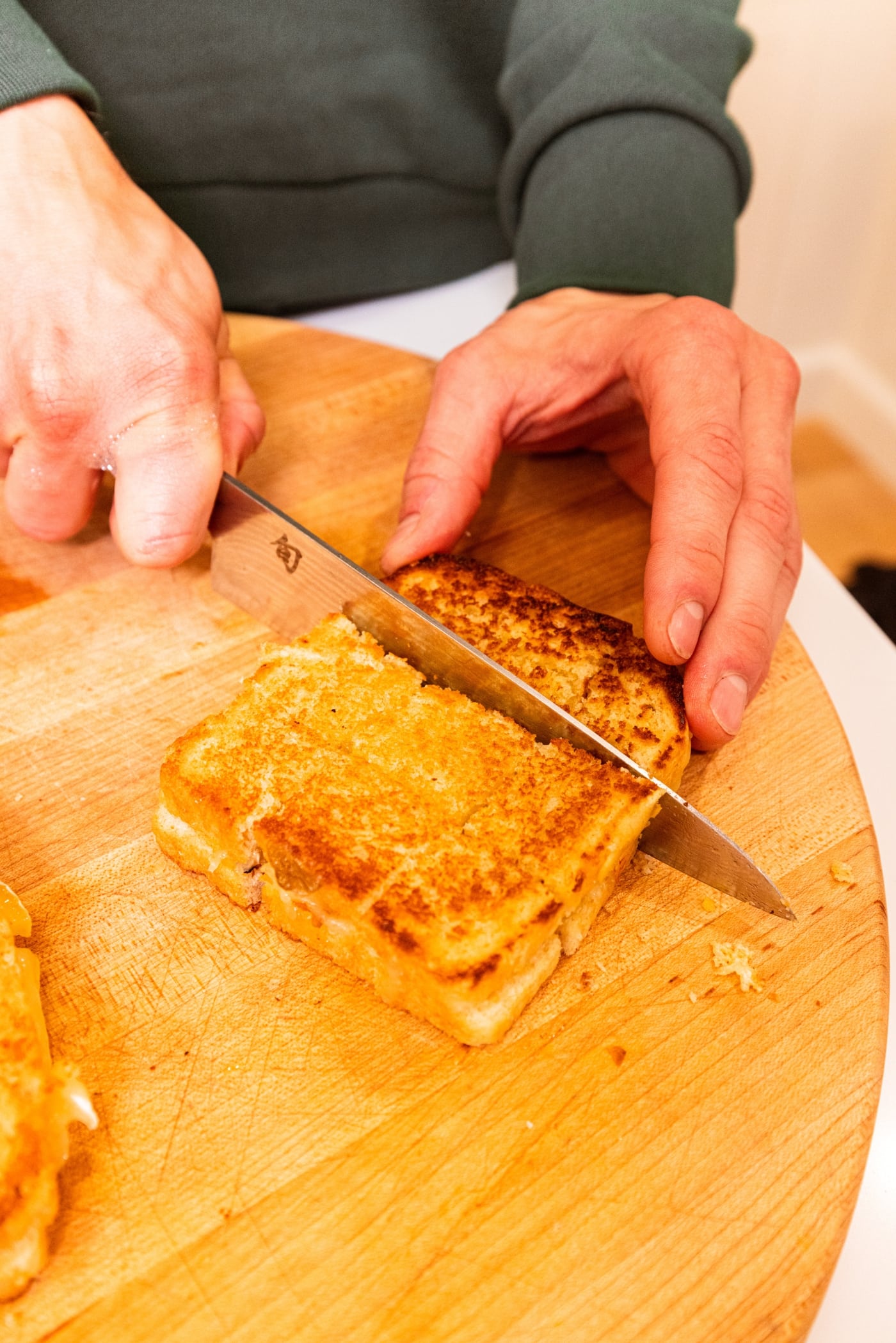 Hand slicing grilled cheese sandwich into nine pieces, equating to bite-sized croutons on wooden cutting board.