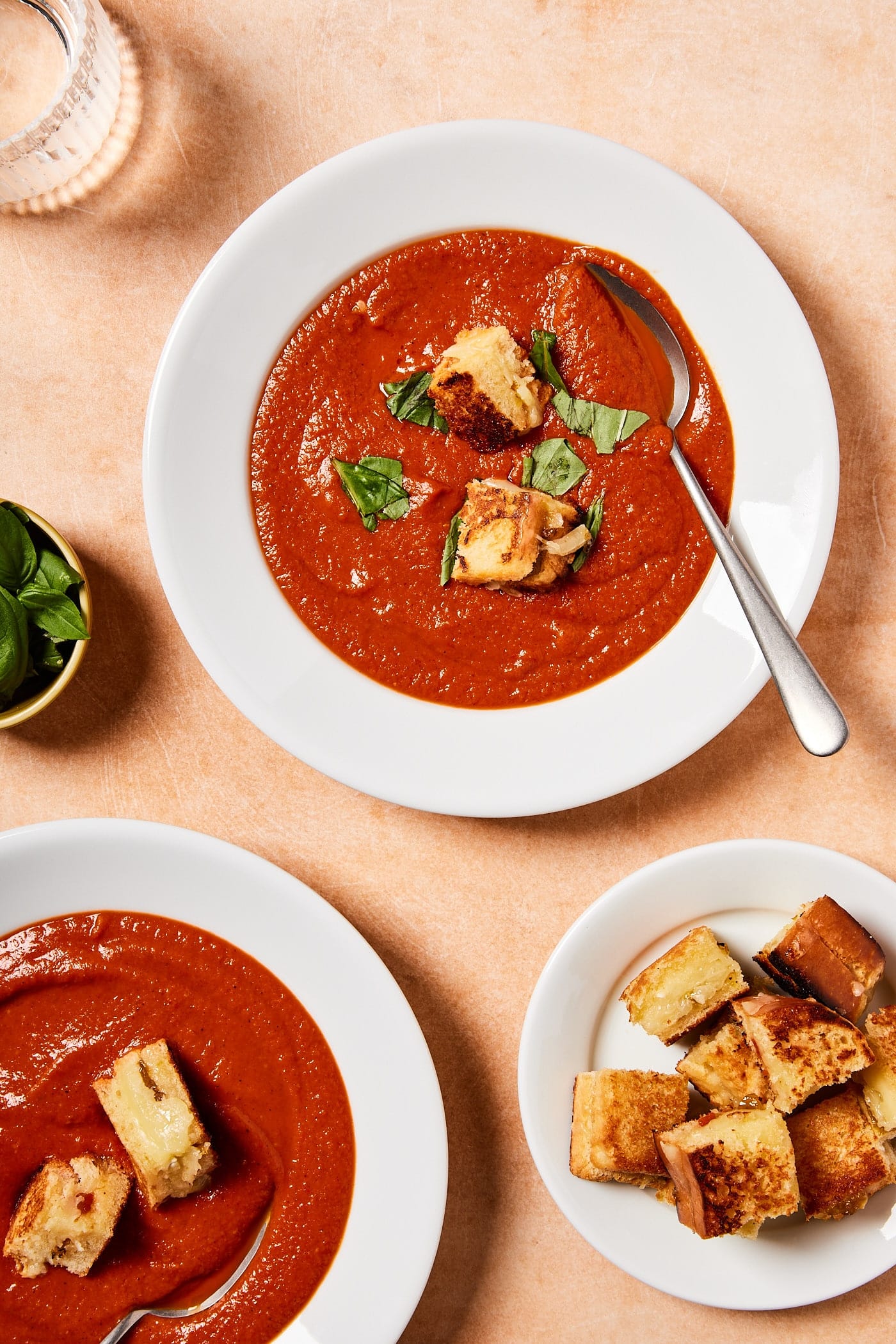 Two white bowls filled with roasted tomato soup with plate filled with grilled cheese croutons to the side.