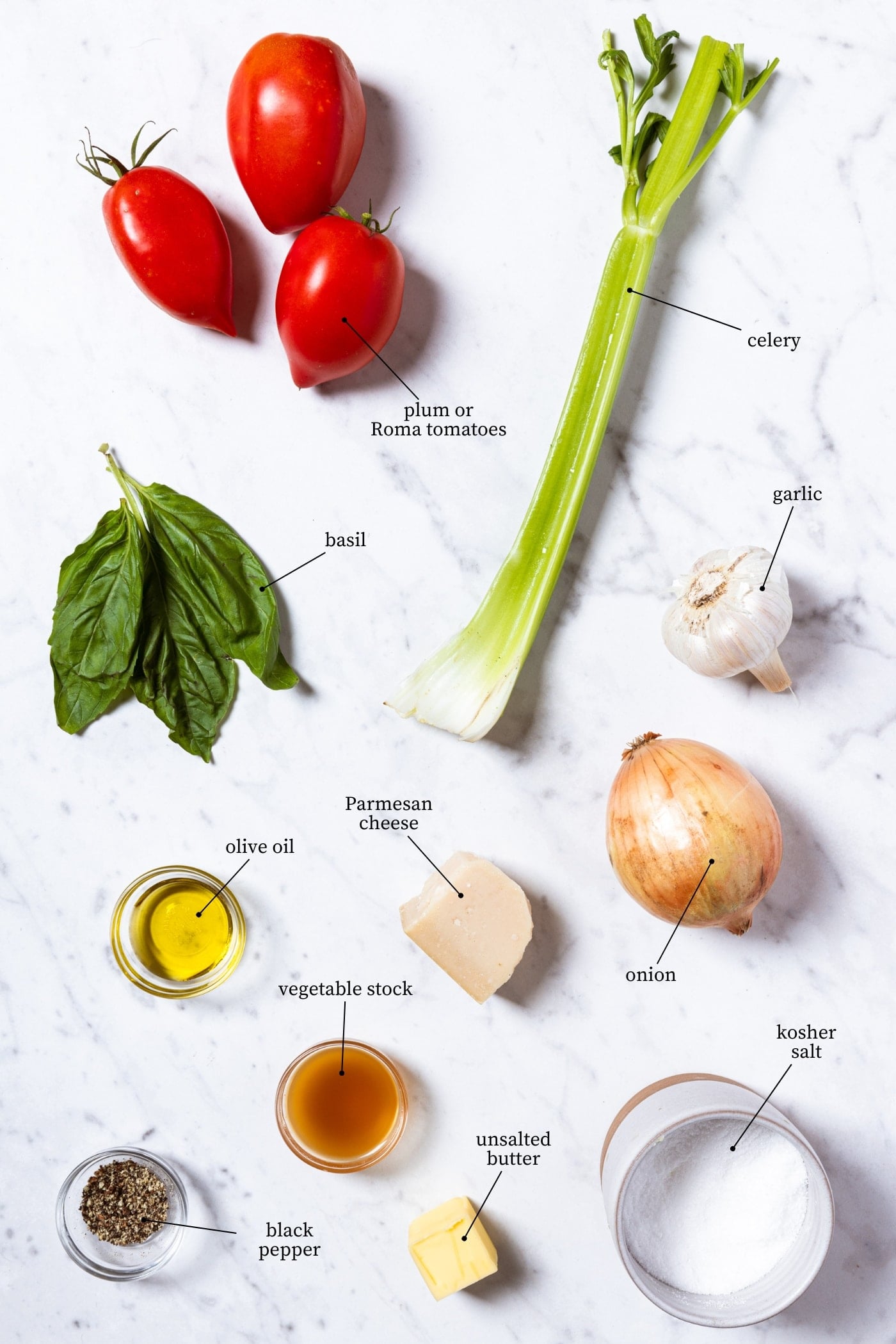 White marble surface filled with all ingredients needed to make roasted tomato soup including tomatoes, celery, basil, spices, and more.