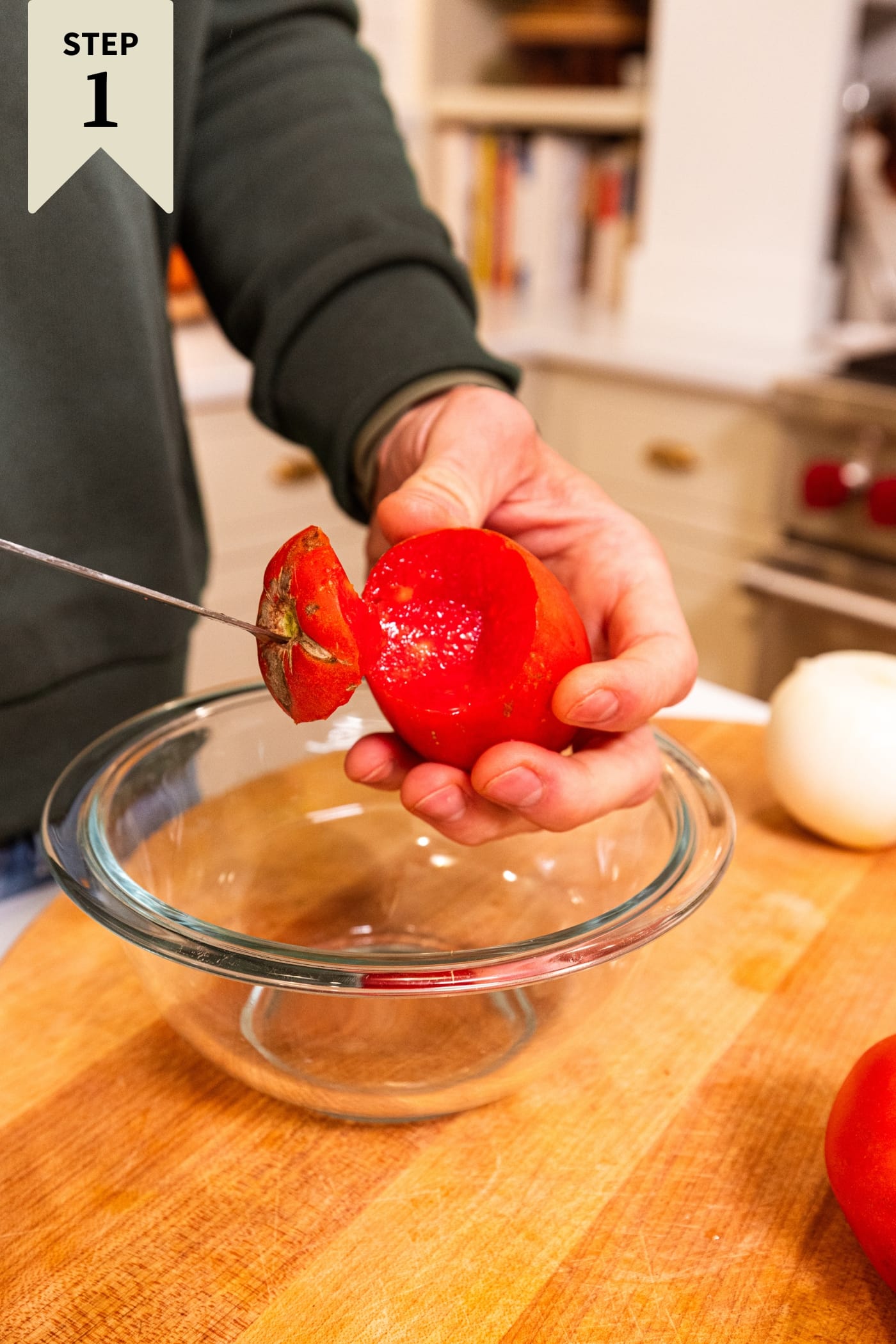 Hand holding knife cutting out the top of a tomato into a glass bowl on wood cutting board.