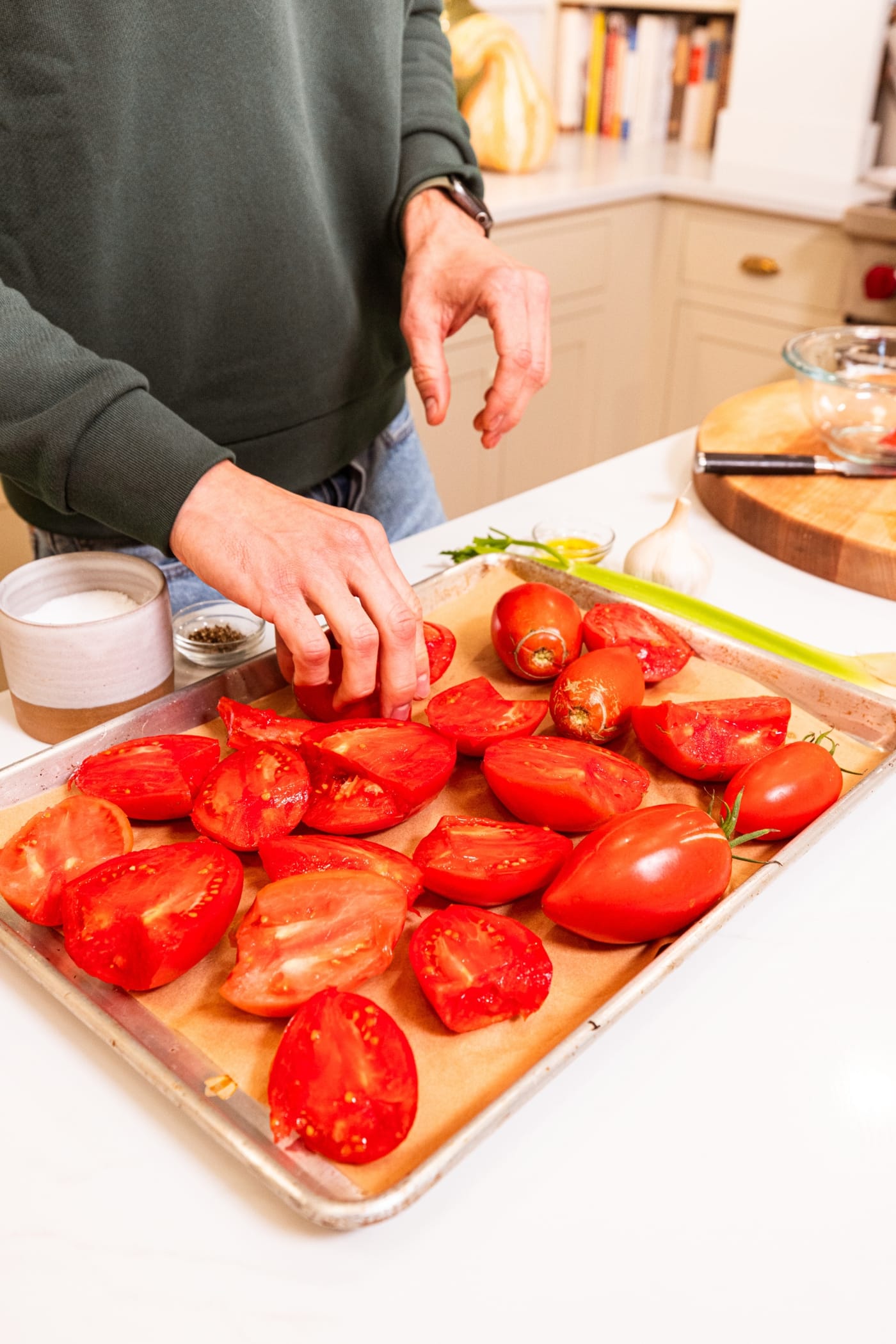Hand placing sliced tomatoes onto a parchment-lined baking sheet with celery and salt and pepper in background.