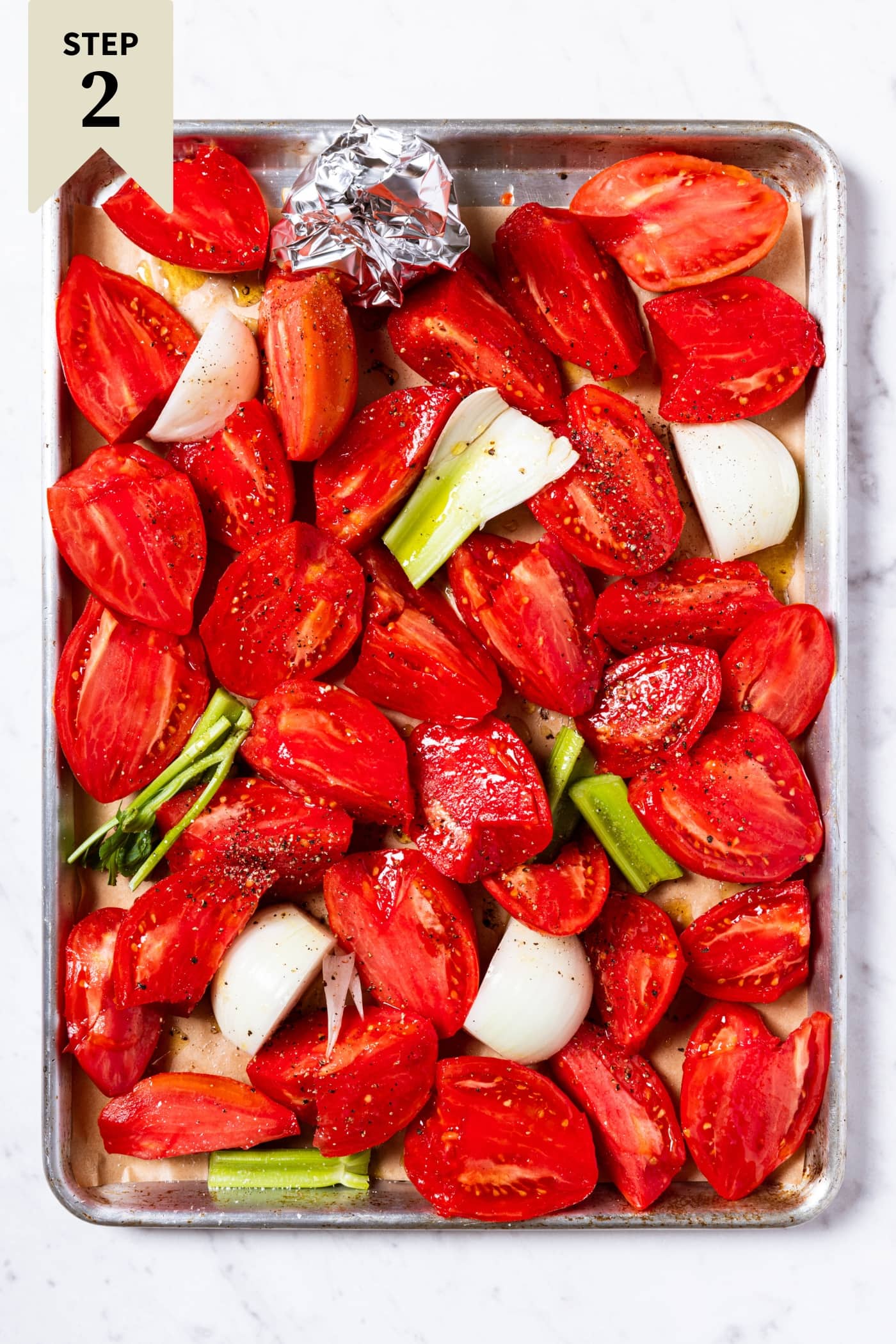 Large baking sheet filled with sliced tomatoes, onions, and celery on white marble surface before going into the oven to bake.