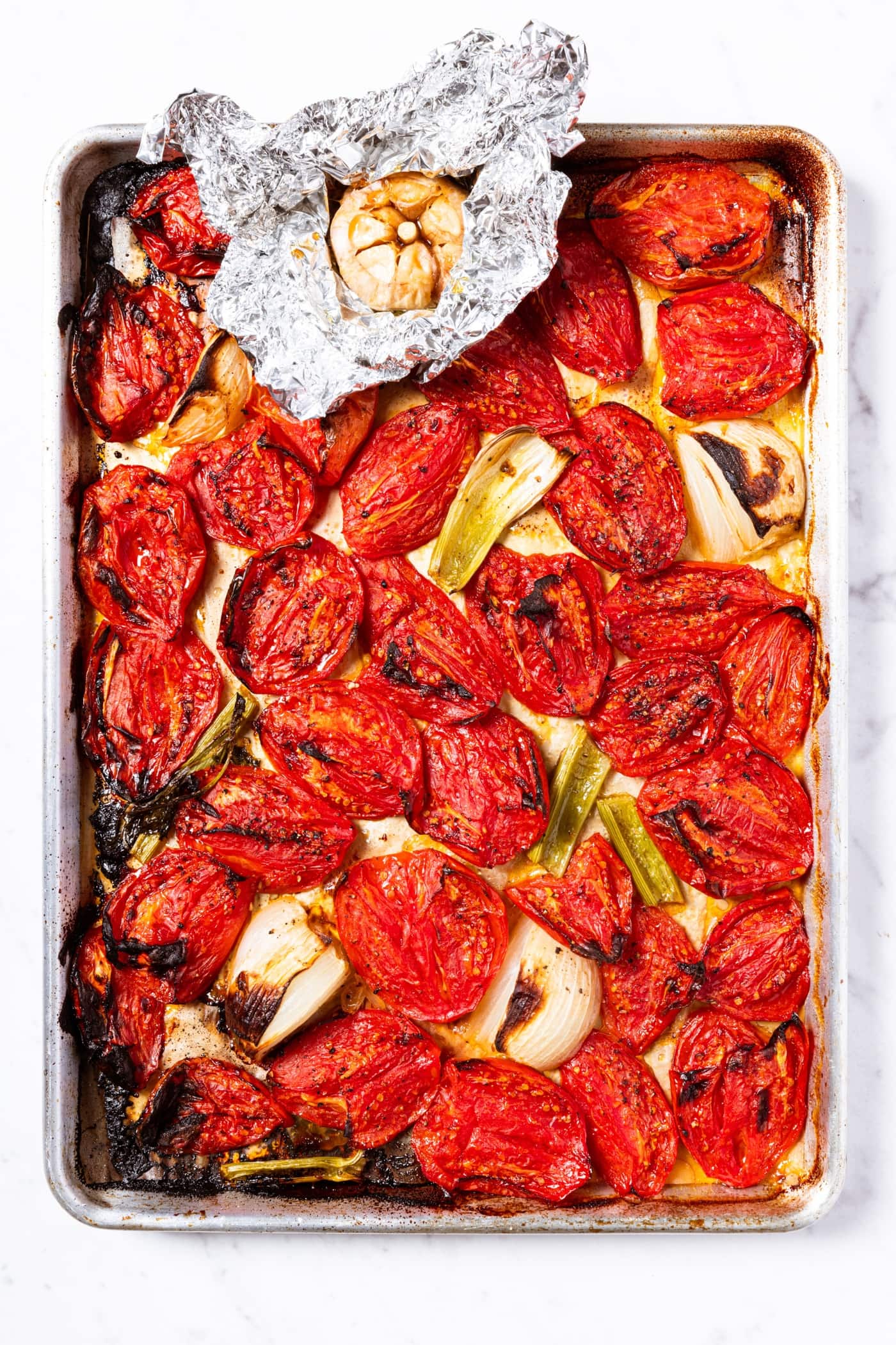 Large baking sheet with roasted tomatoes and other vegetables and garlic clove in tin foil.