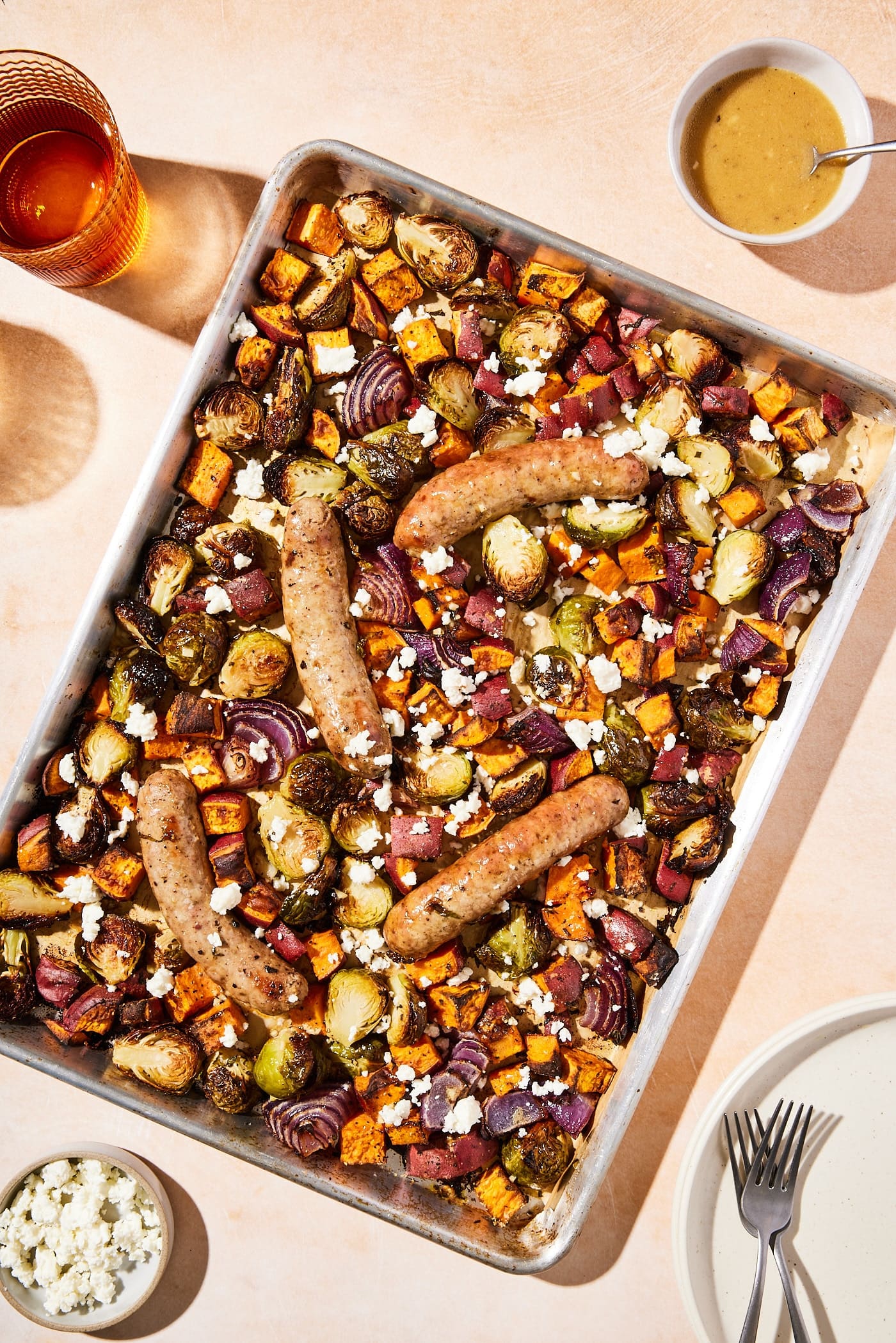 Large metal sheet pan with roasted vegetables and sausage with glasses and plates.