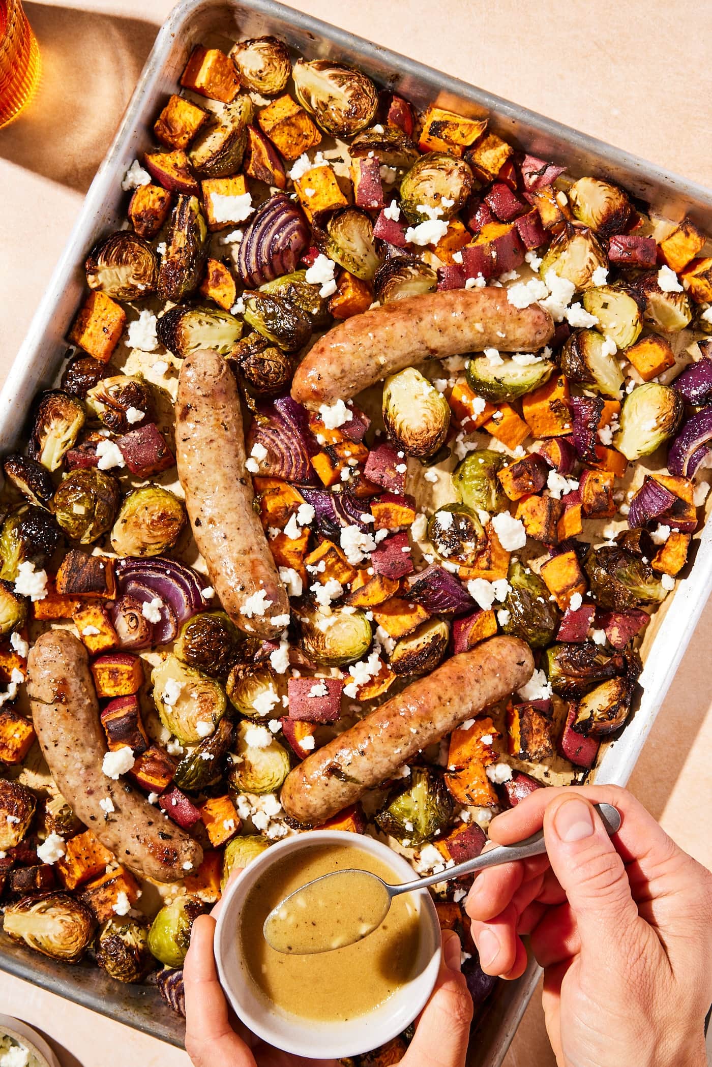 Hand holding spoon about to drizzle vinaigrette on top of roasted vegetables and sausage on a sheet pan.