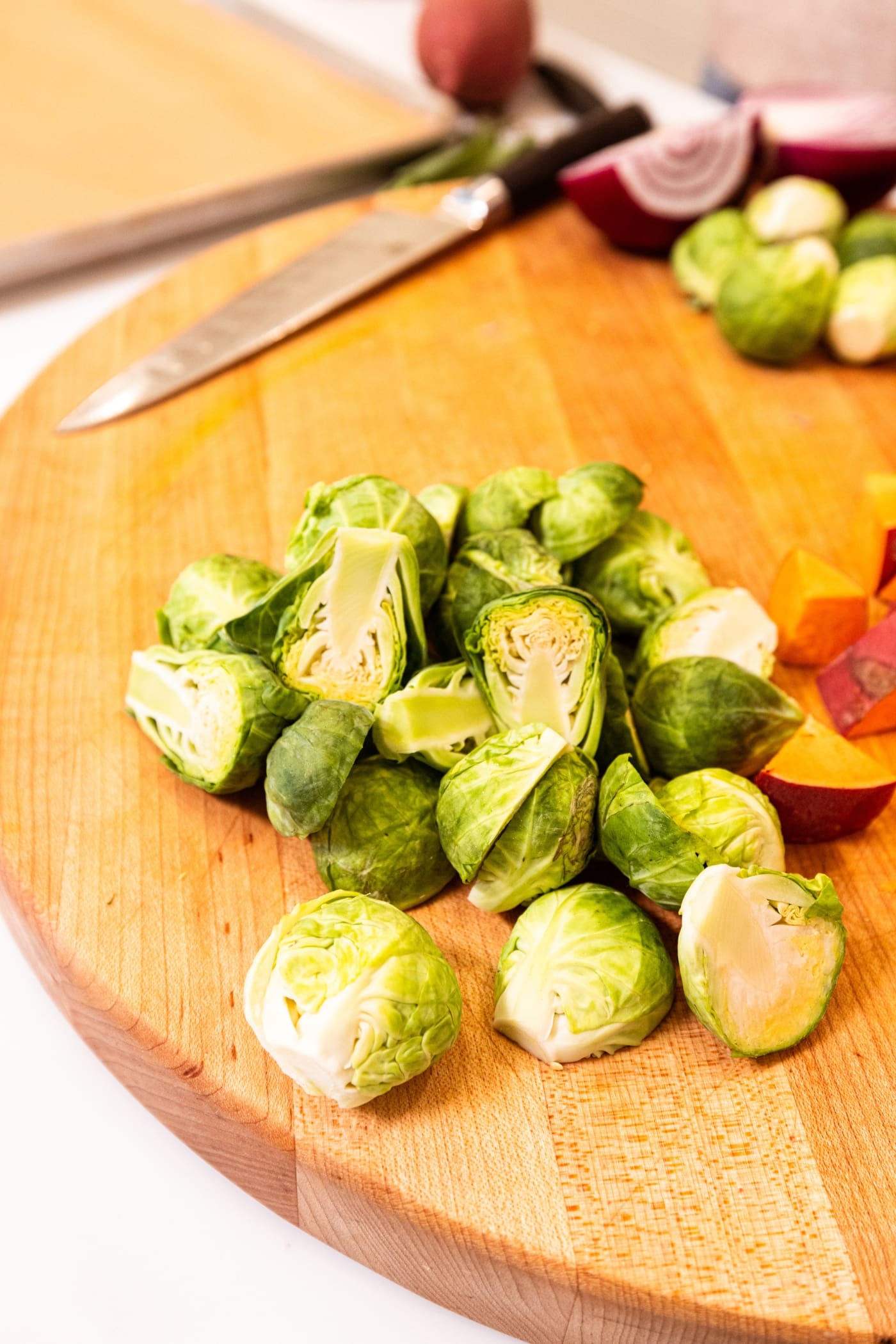 Sliced Brussels sprouts sitting on a wood cutting board with chopped sweet potatoes to the side.
