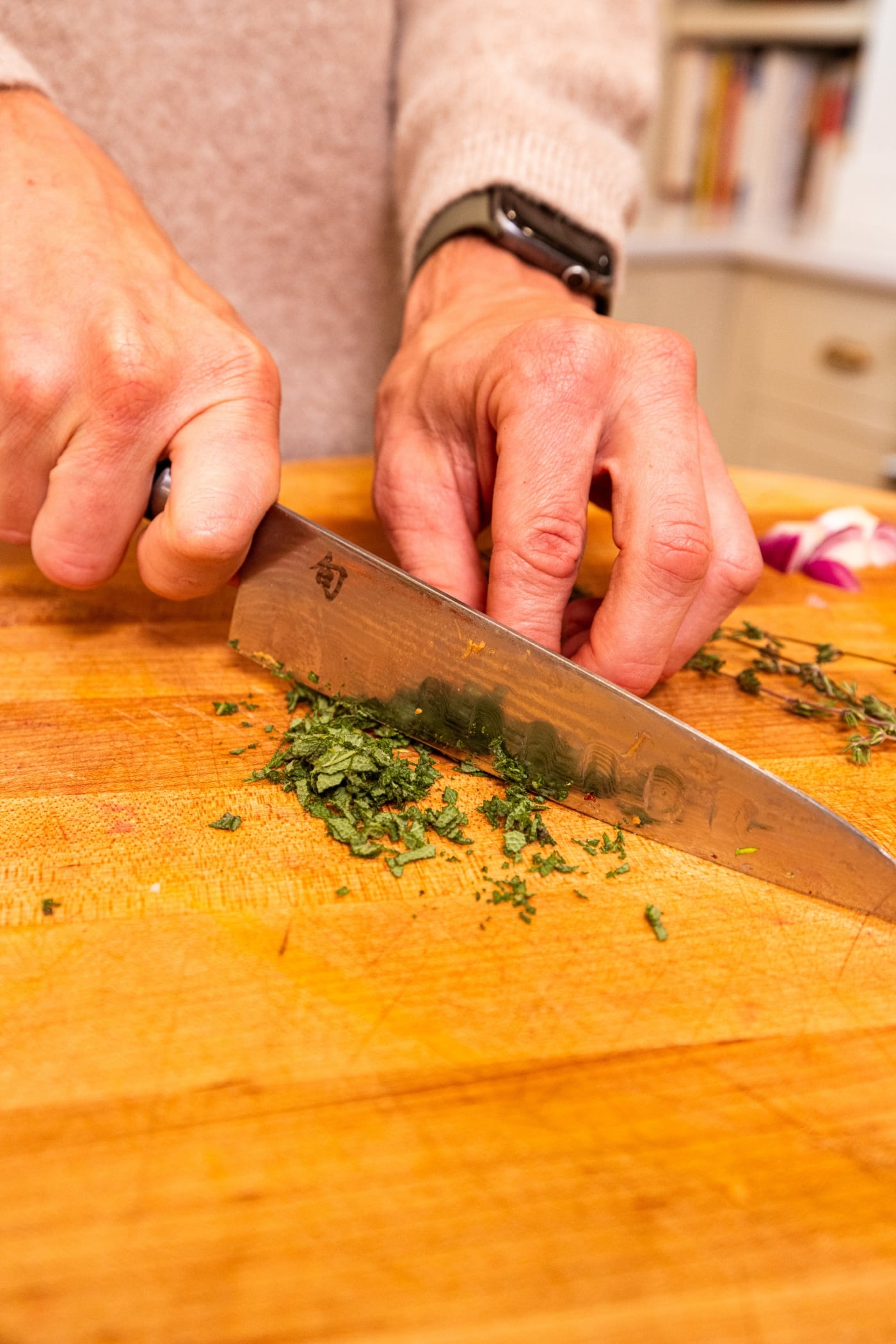 Two hands using a knife to chop up fresh herbs on a wooden cutting board.