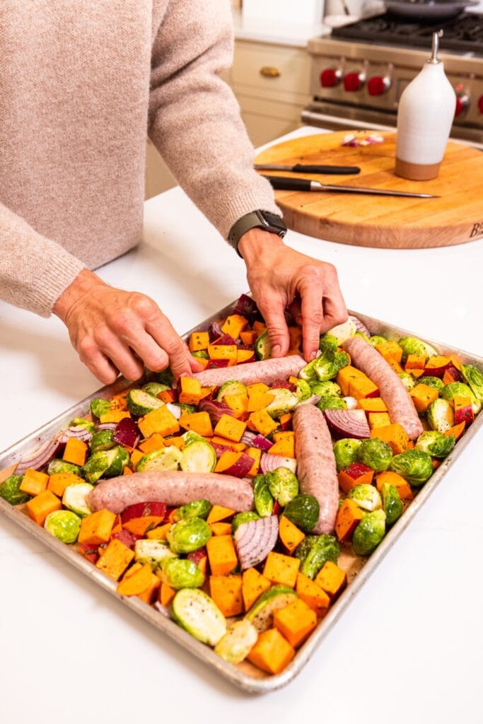 Two hands nestling raw sausages among raw chopped vegetables on a sheet pan.
