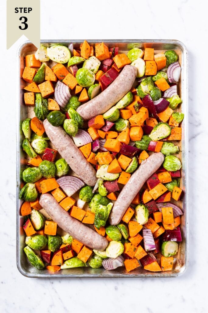 Sheet pan on a marble surface with raw vegetables and raw sausage before going into the oven to roast.