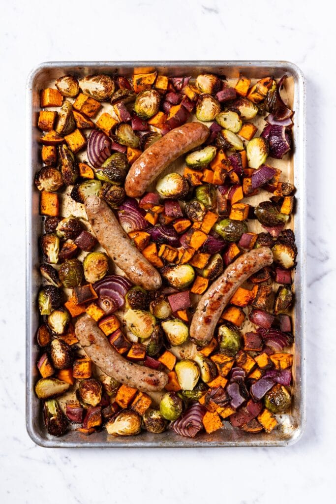 Sheet pan on marble surface with cooked vegetables and sausage after roasting in the oven.