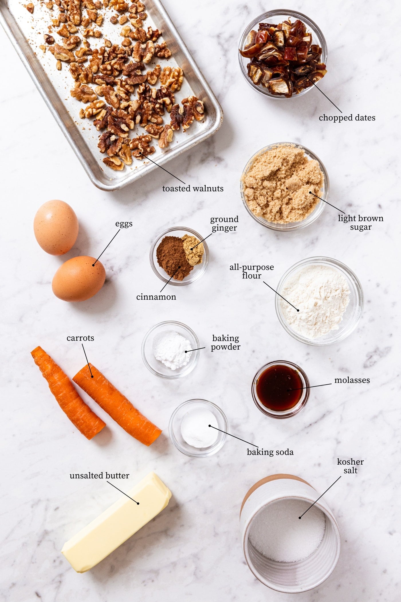 White marble surface filled with all ingredients needed to make spiced carrot muffins including raw carrot, walnuts, eggs, spices, and more.