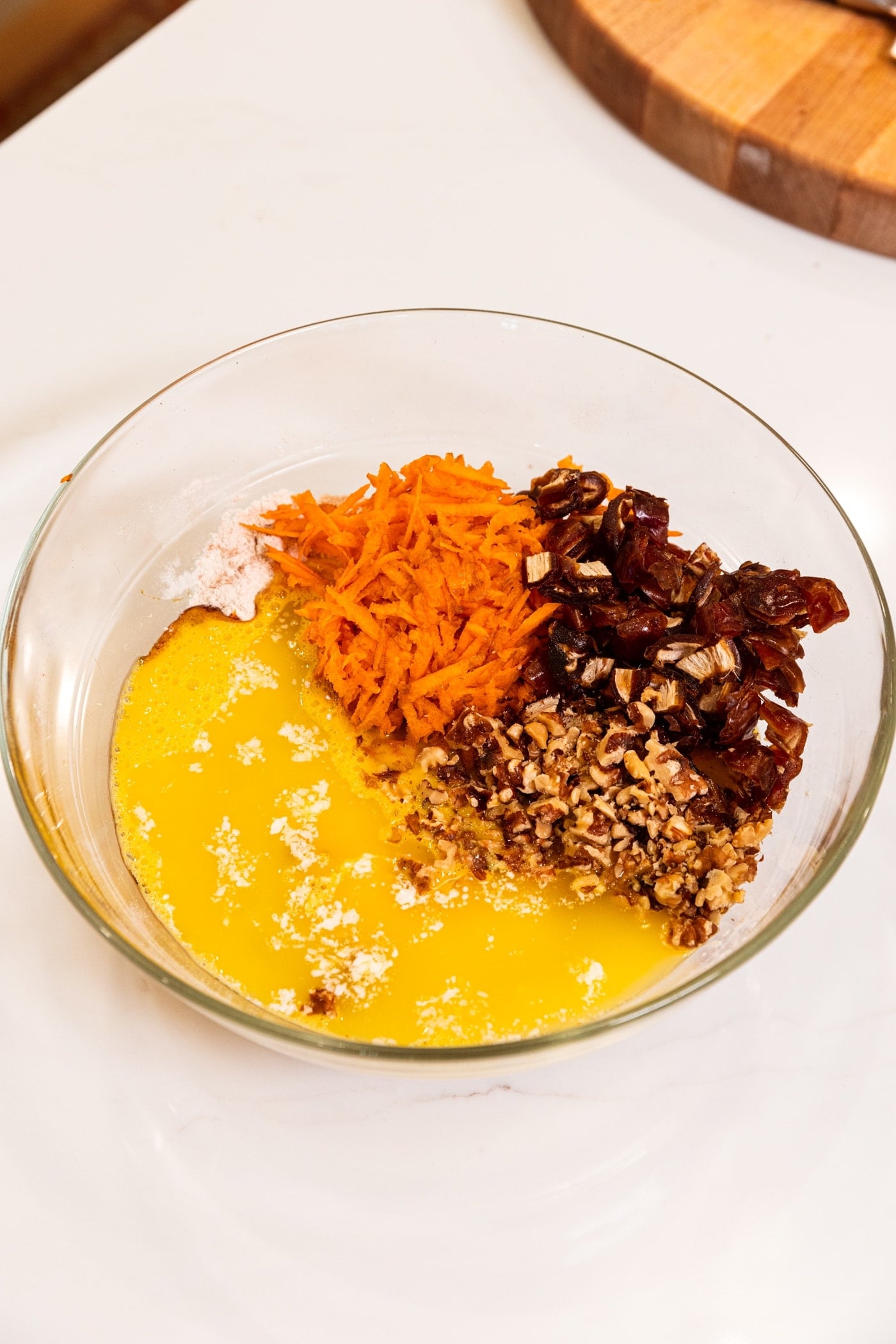 Glass bowl filled with wet ingredients for carrot muffins including shredded carrot, dates, nuts, and eggs.