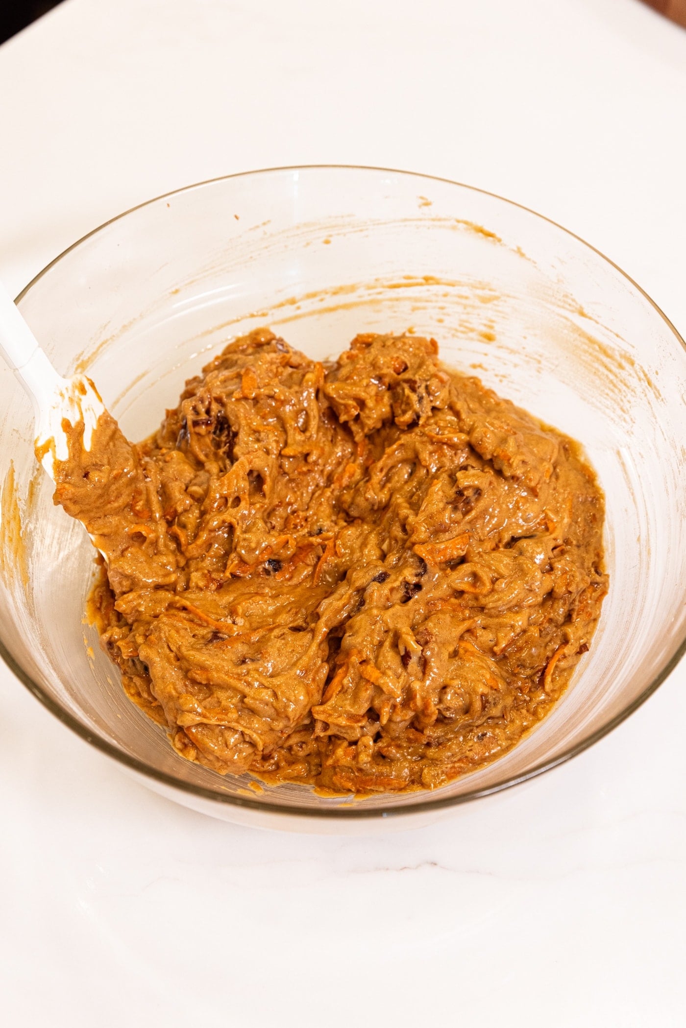 Plastic spatula resting in glass bowl with batter ready for spiced carrot muffins.