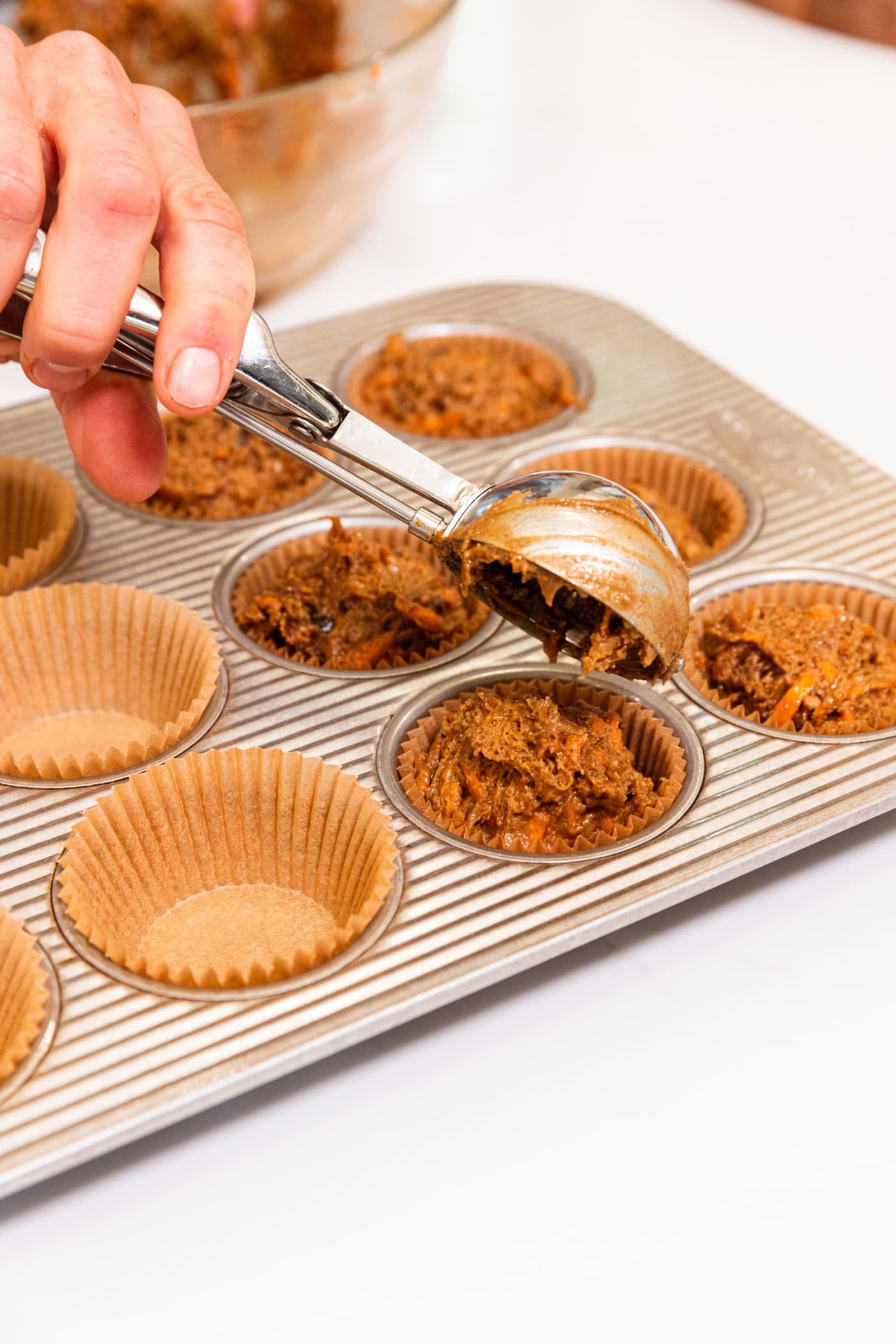 Hand using cookie scoop to place dollops of batter into a muffin pan fitted with muffin cups.