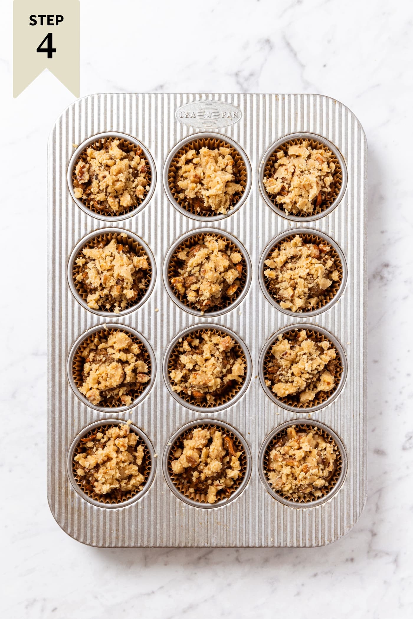 Muffin tin pan resting on white marble surface filled with unbaked carrot muffins with streusel topping.
