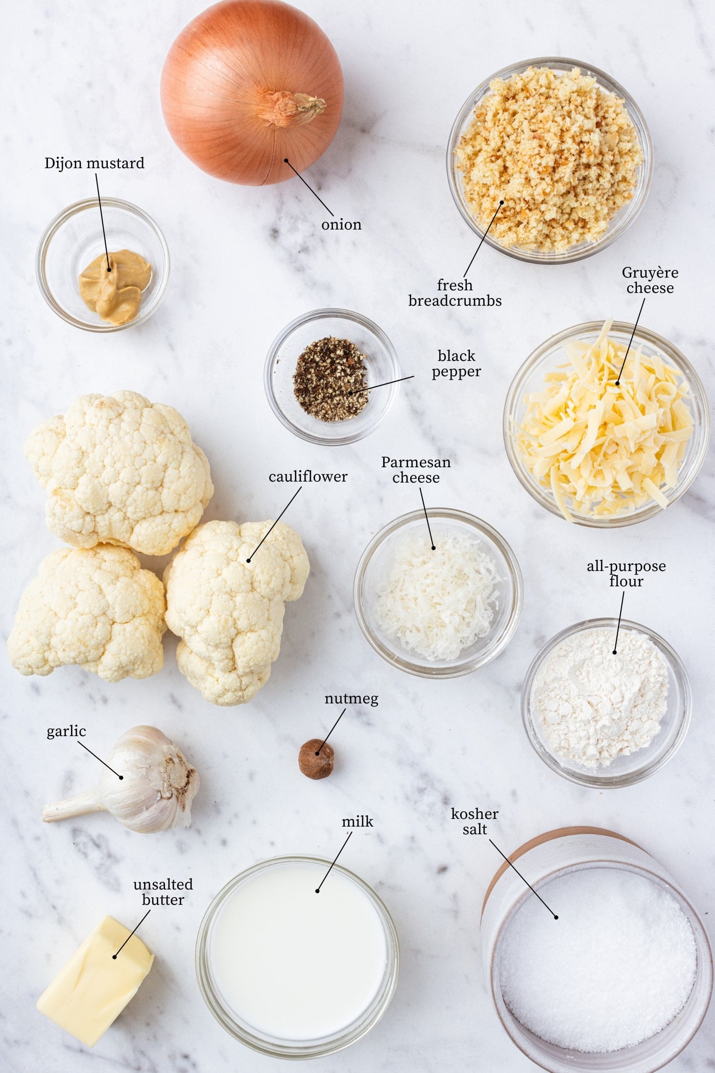 White surface filled with all ingredients needed to make cauliflower au gratin including cauliflower, spices, cheese, milk, breadcrumbs, and more.