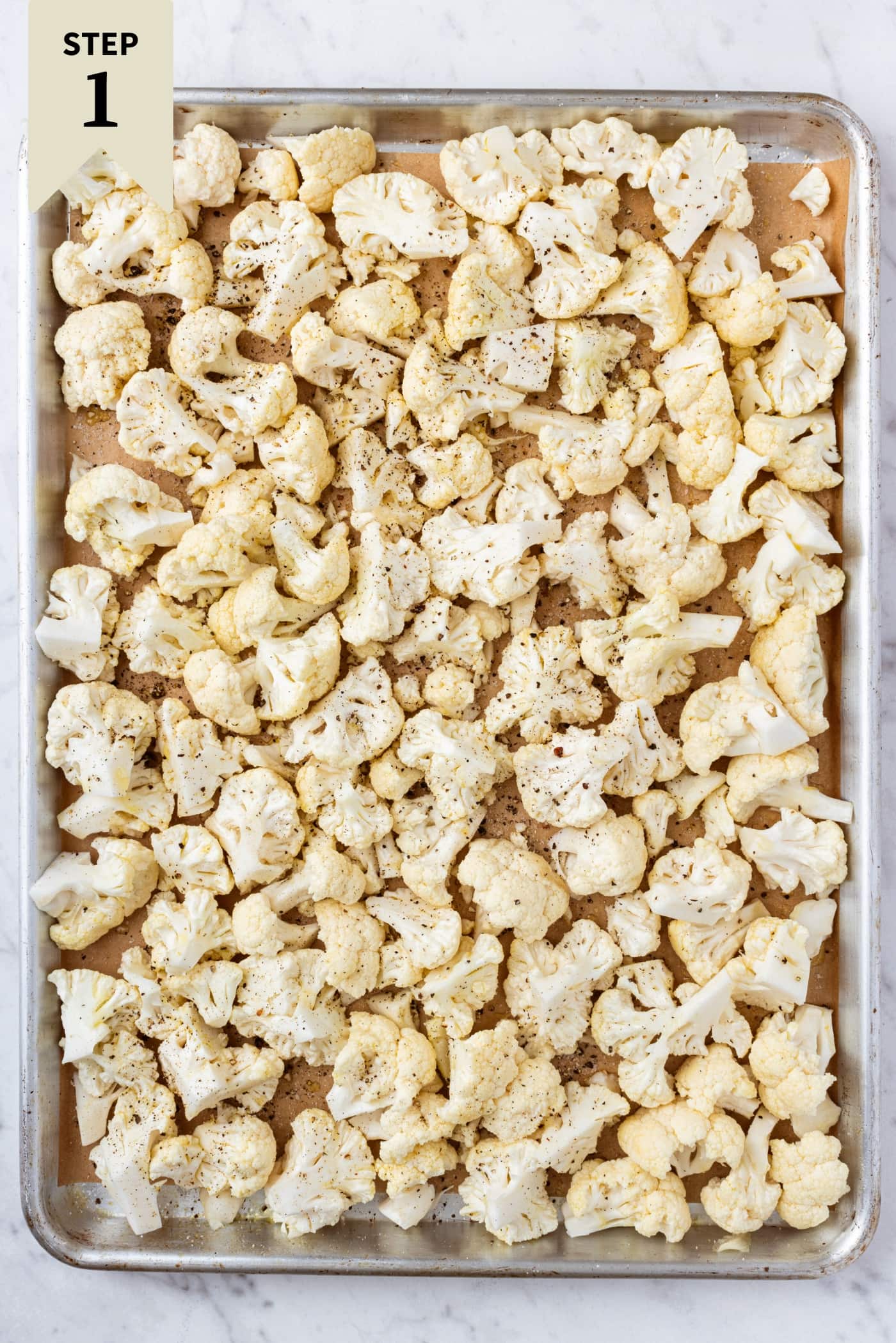 Metal roasting pan filled with raw cauliflower florets drizzled with olive oil and sprinkled with salt and black pepper before going into the oven to roast.