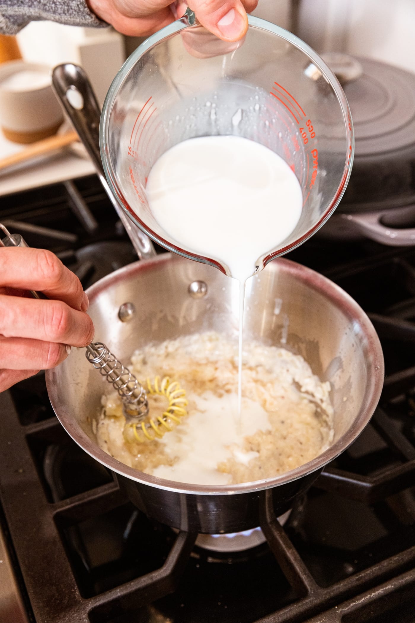 Large measuring cup pouring milk into a metal saucepan saucepan filled with cooked flour mixture.