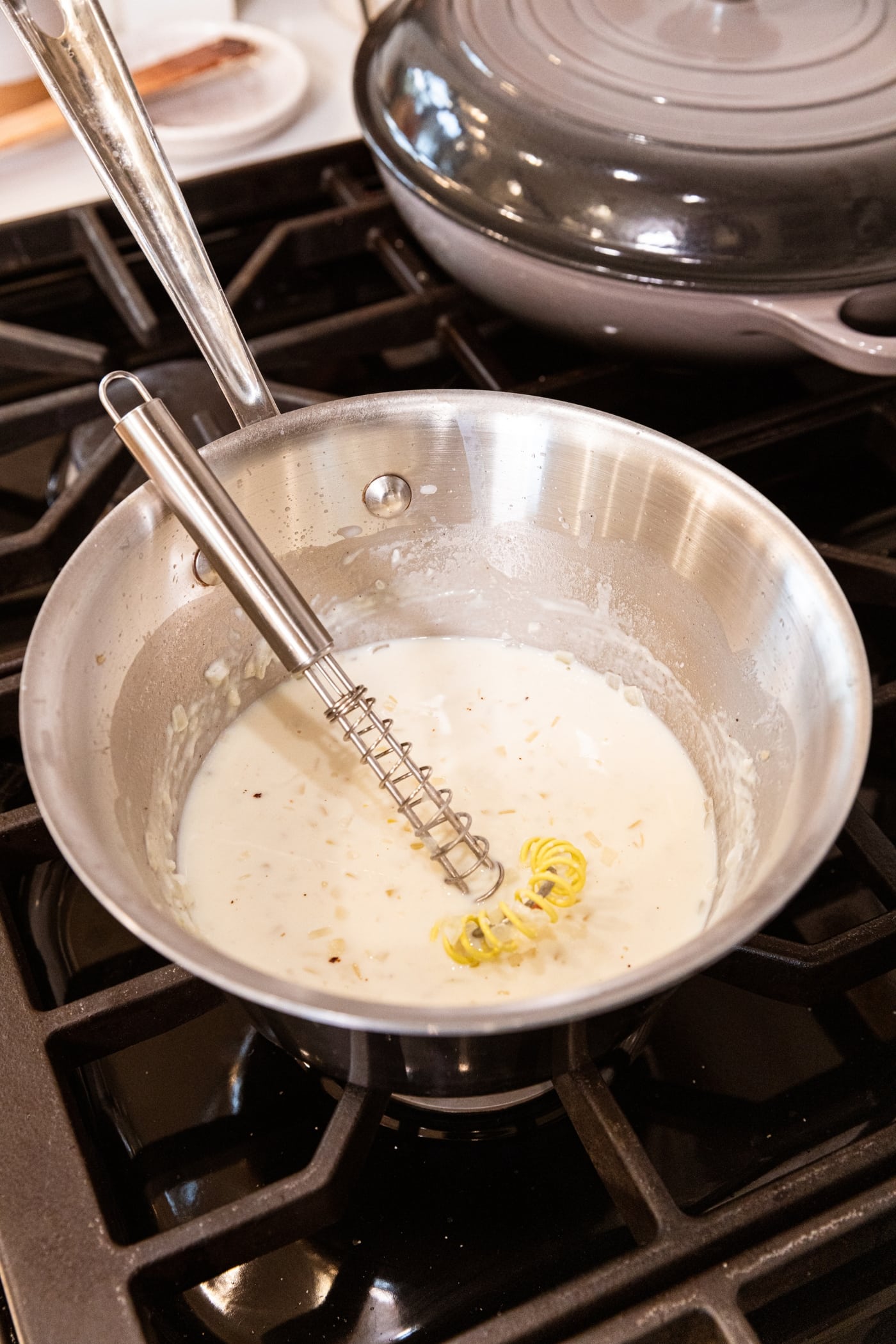 Metal whisk resting against the side of a metal saucepan filled with cooking milk and flour mixture.