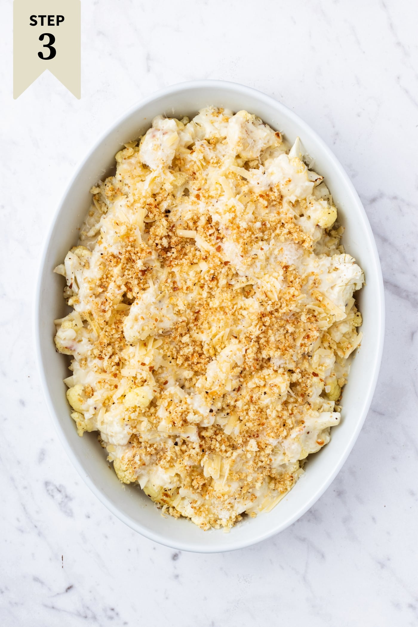 Marble surface with white baking dish filled with unbaked cauliflower au gratin topped with fresh breadcrumbs right before going into the oven.