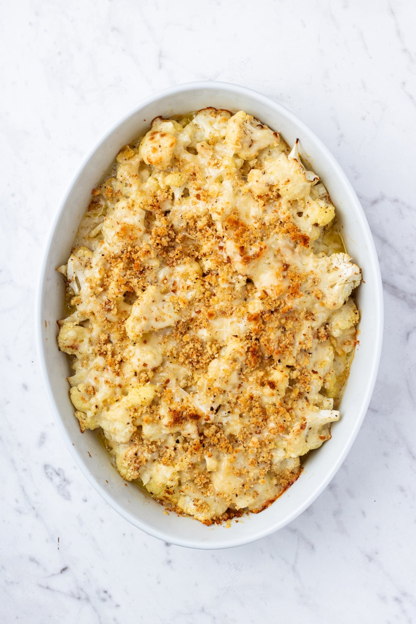 White marble surface with white baking dish filled with cauliflower au gratin topped with fresh breadcrumbs right after coming out of the oven.