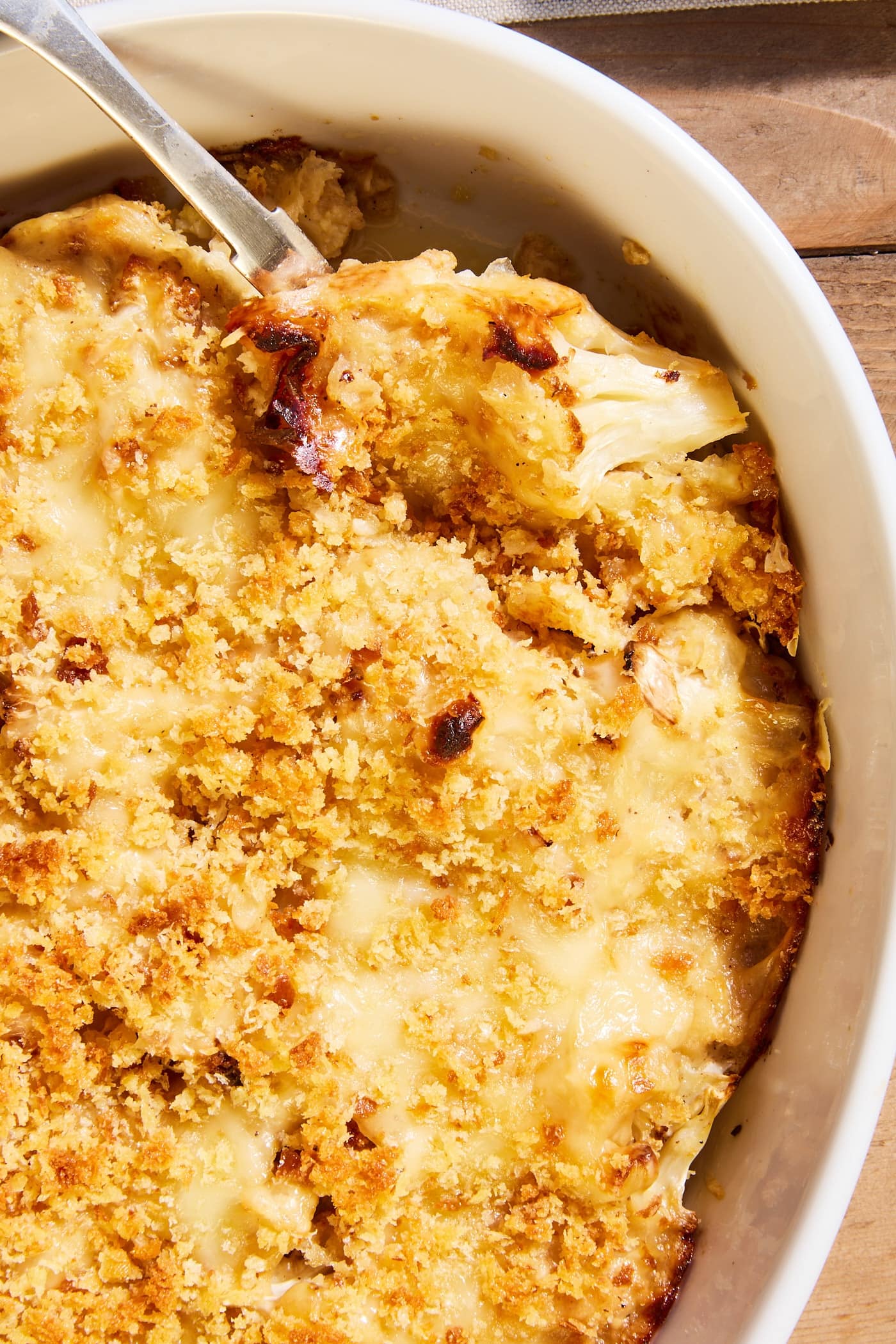 White baking dish filled with baked cauliflower au gratin with crispy golden brown topping with spoon scooping out a serving.