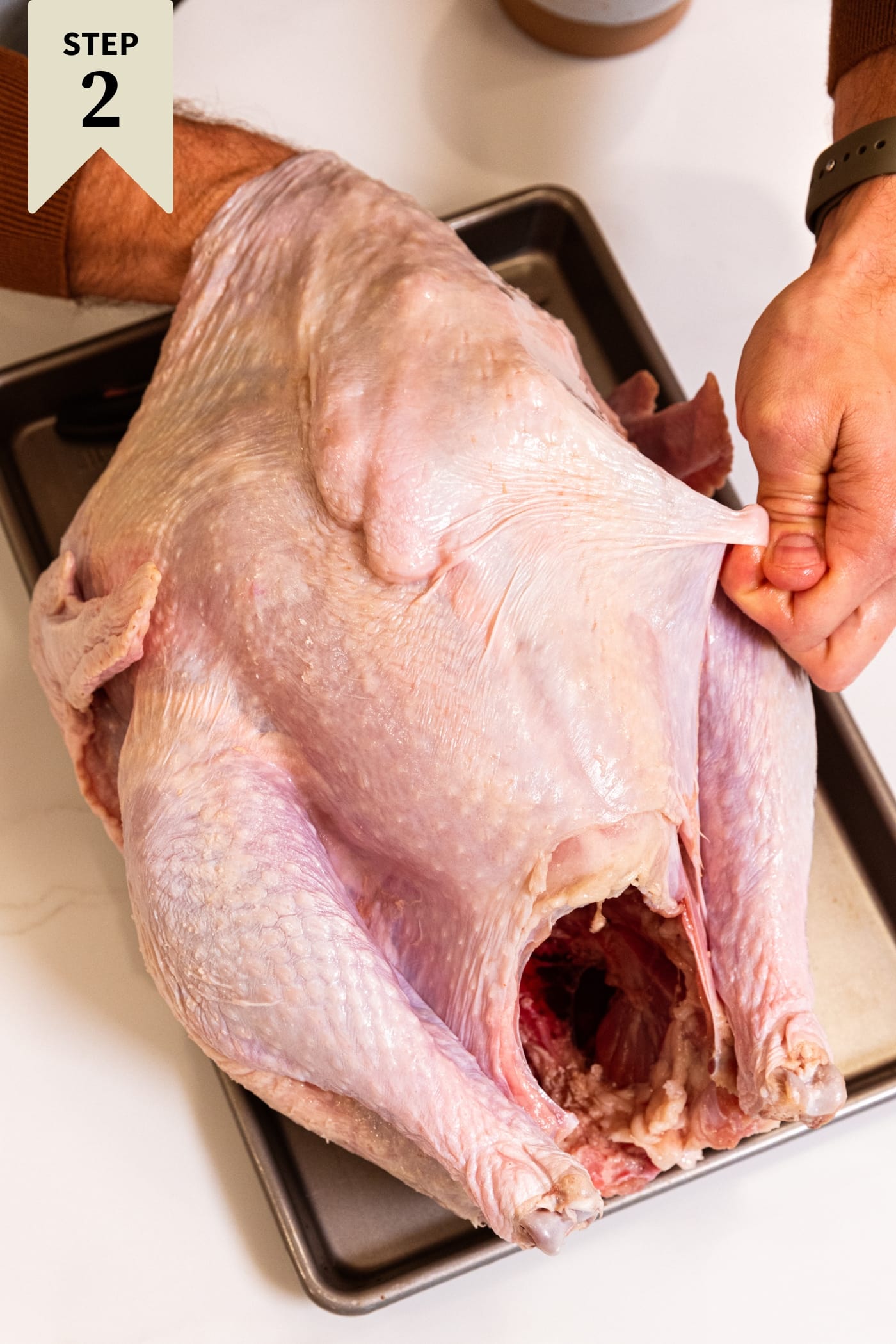 Two hands pulling away the skin from a raw turkey on a metal pan.