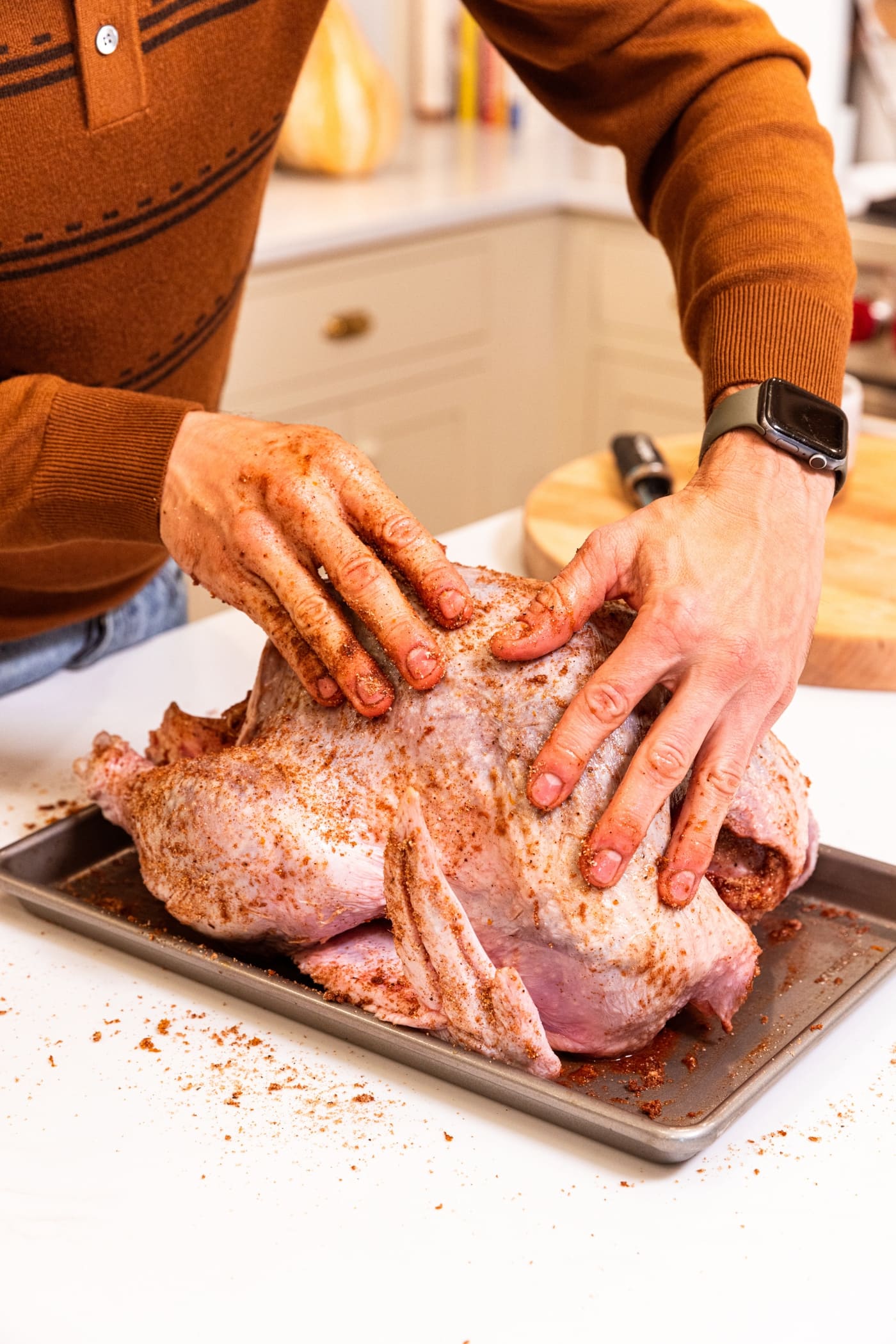 Two hands brushing brown colored dry brine all over the top of a raw Thanksgiving turkey on a metal pan.